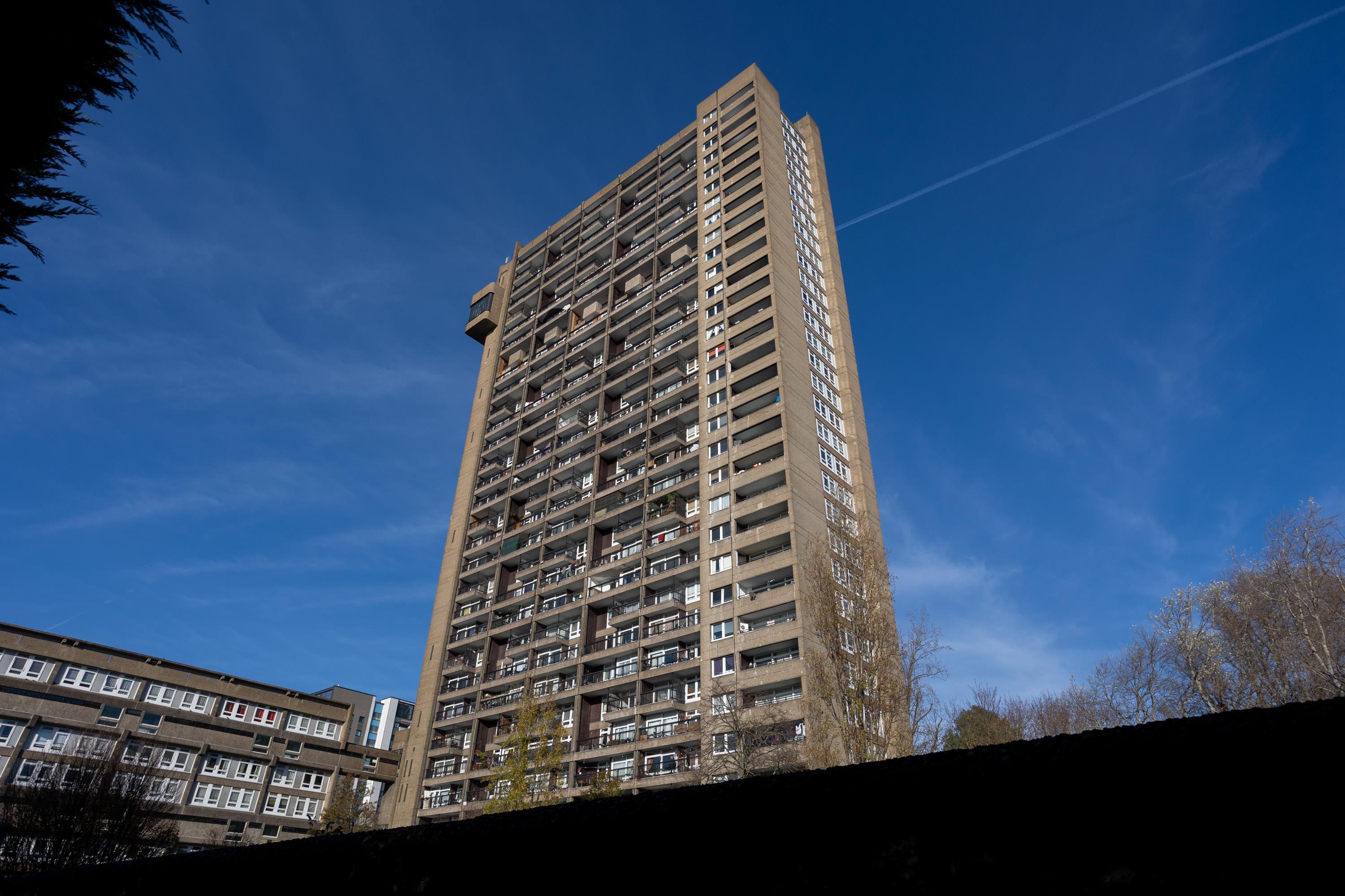 A tall, rectangular concrete block of flats with many balconies stands against a blue sky. Shorter buildings and leafless trees are visible at the base. Contrail lines are in the sky. The scene is well-lit by sunlight.