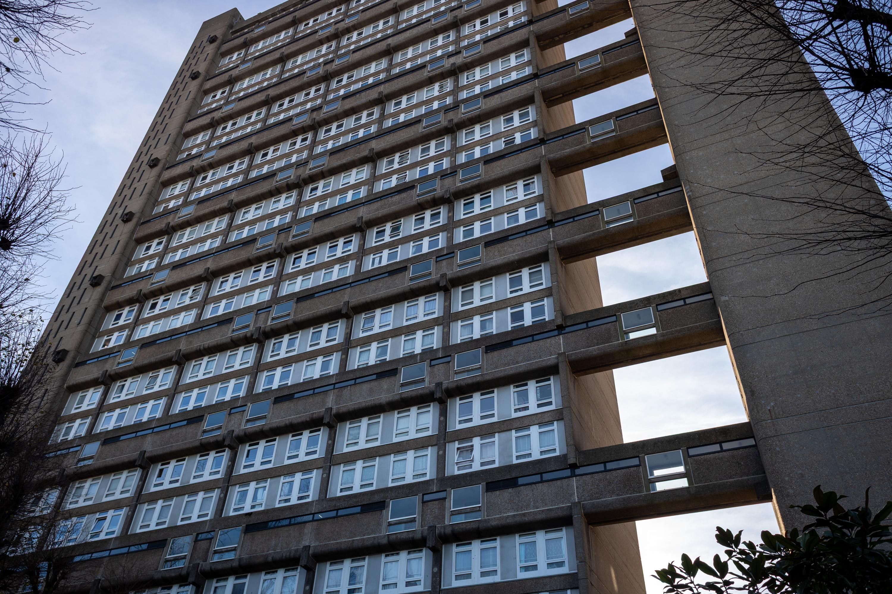 A tall, brutalist block of flats with many rows of white-framed windows and concrete balconies, viewed from below. The sky is partly visible, and leafless tree branches frame the scene on both sides.