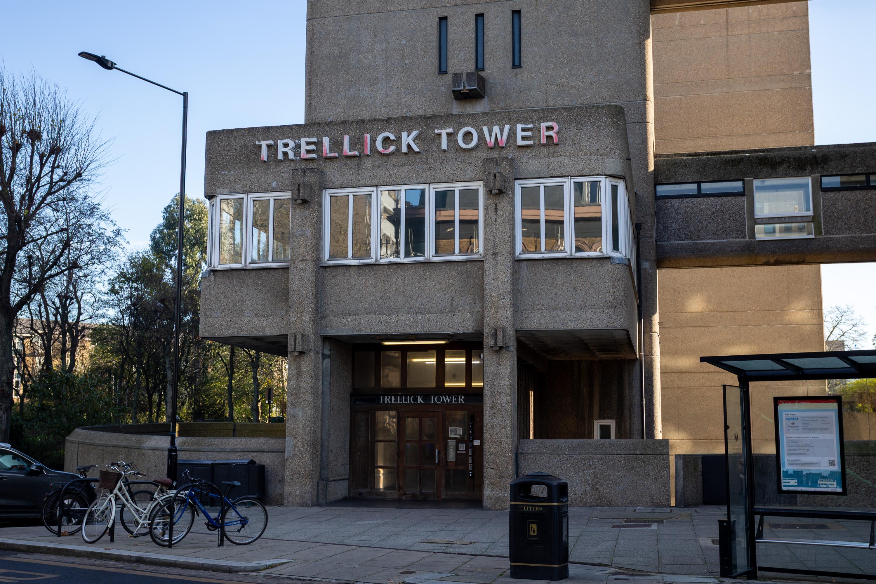 The entrance to Trellick Tower, a concrete residential building, with large block letters above the doors. Three bicycles are parked nearby, and a bus stop stands to the right with a timetable display. Trees and blue sky are visible in the background.