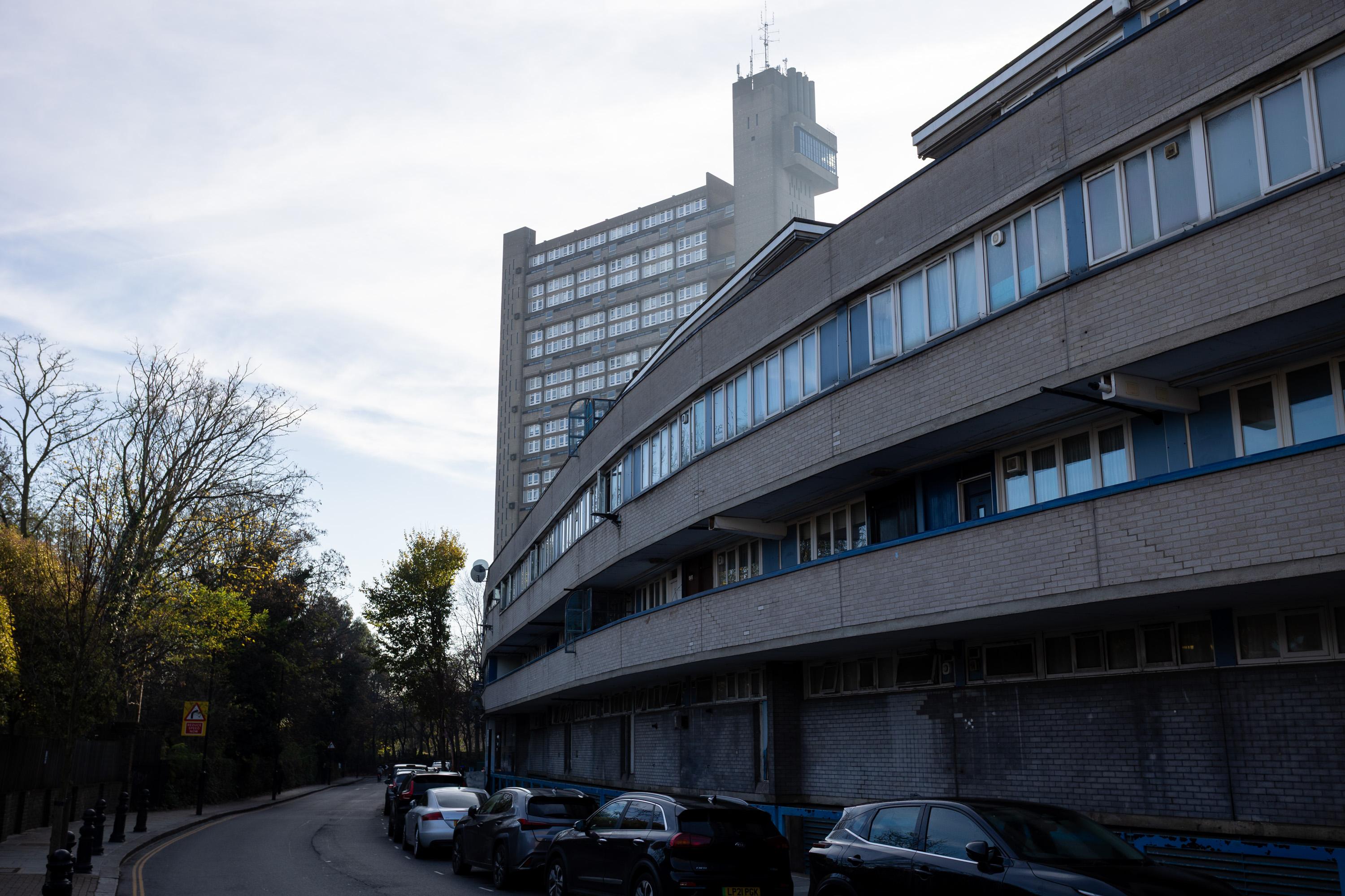 A curved, grey, multi-storey block of flats lines the right side of a street with parked cars. In the background, a tall tower block rises against a bright, partly cloudy sky. Trees with sparse leaves stand to the left of the scene.