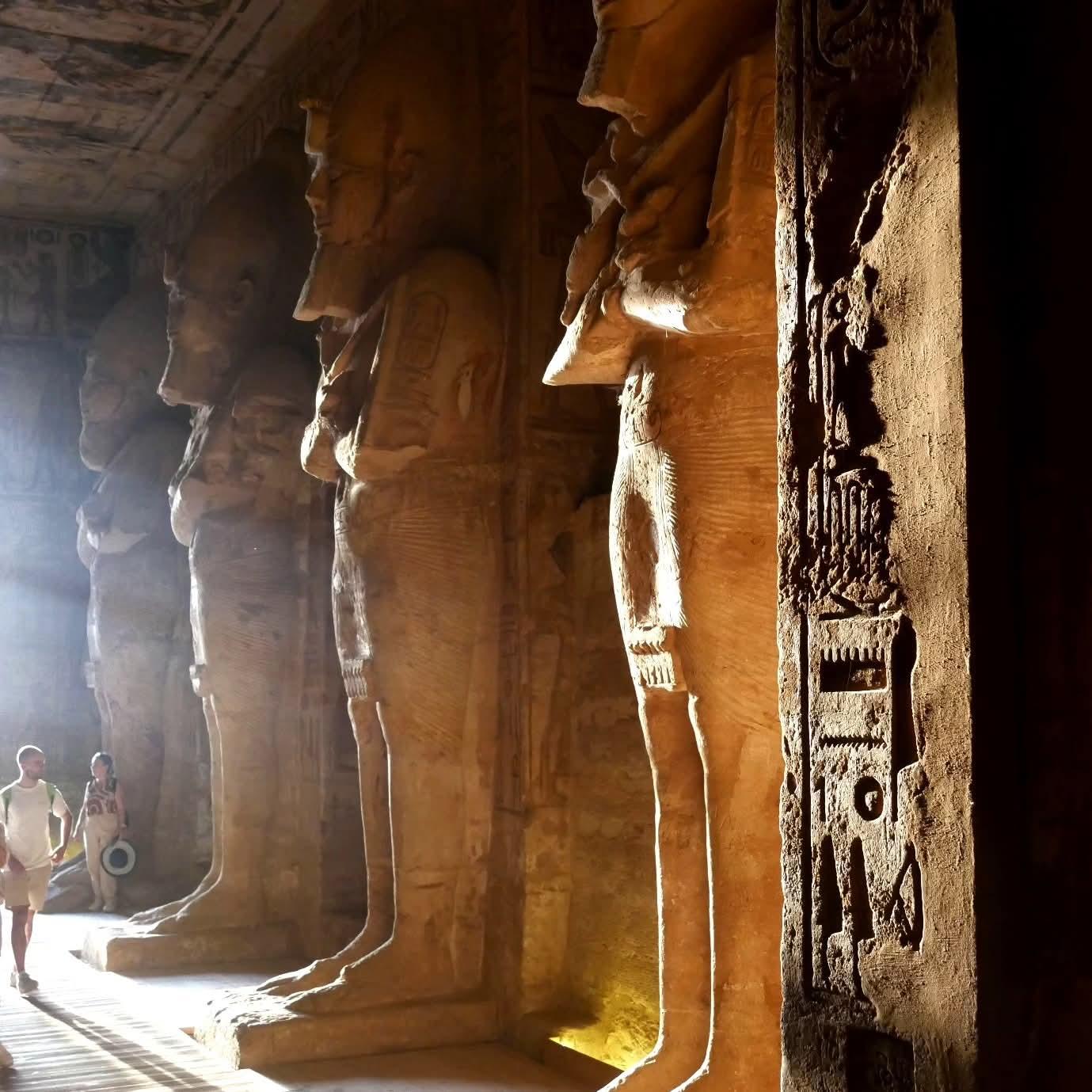 Innenseite des Tempel von Ramses II in Abu Simbel. Die Morgensonne dringt in den heiligen Tempel und erfüllt alles in einen goldenen Glanz. Es sind Statuen des Pharao und Hieroglyphen an der Wand erkennbar.  Besucher betreten gerade das Heiligtum. Ägypten 2023