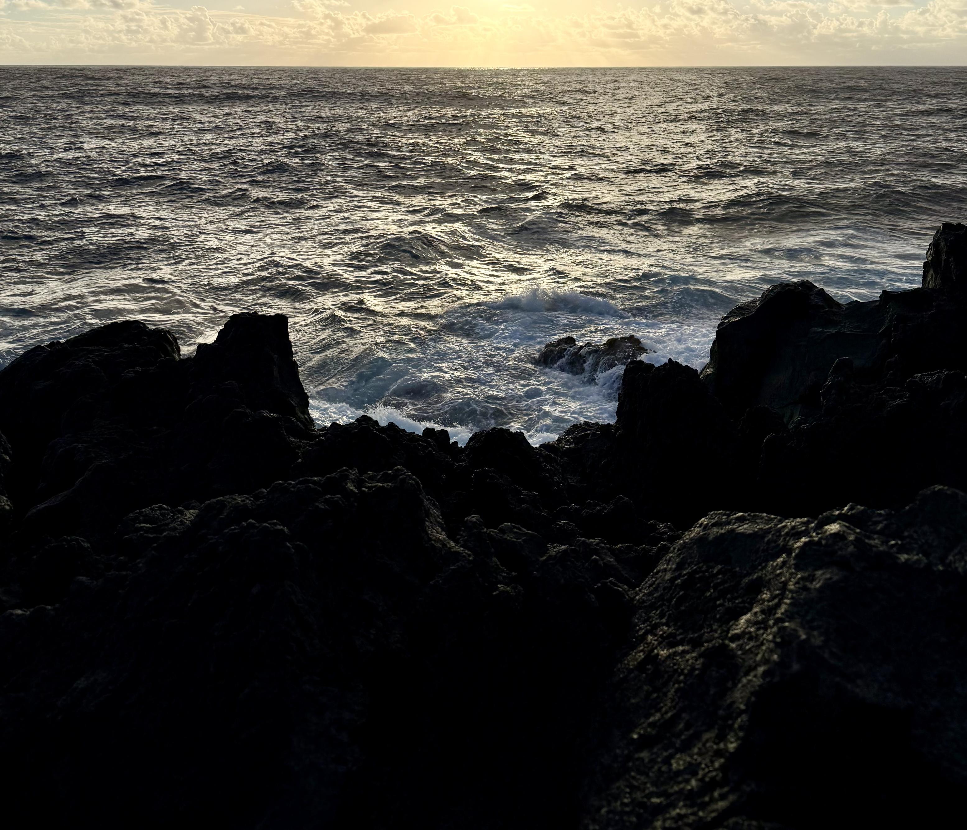 Moody photo shot over jagged deep black lava rocks in the bottom third of the image to a roiling steely gray ocean that’s reflecting the light of the glowing sun just rising over the horizon in a thin strip of ethereally pale cloudy sky at the top.