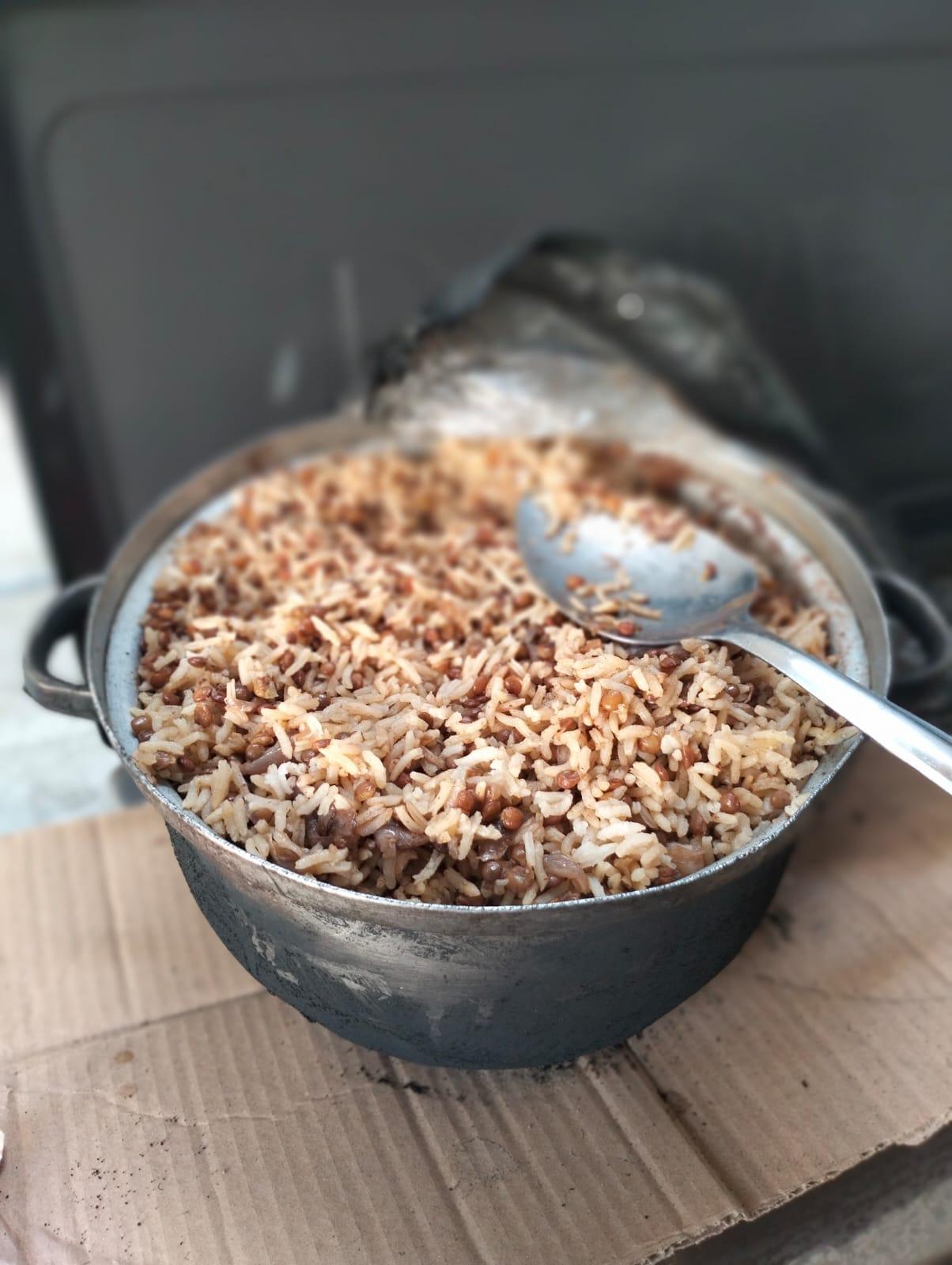 A large pot of cooked rice and lentils with a serving spoon rests on a piece of cardboard.