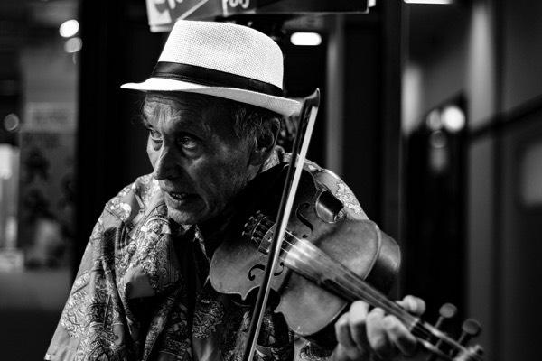 Black-and-white close-up of an older man playing a violin, wearing a light fedora with a dark band and a patterned shirt, focused with an expressive, open-mouthed expression as he draws the bow across the strings.