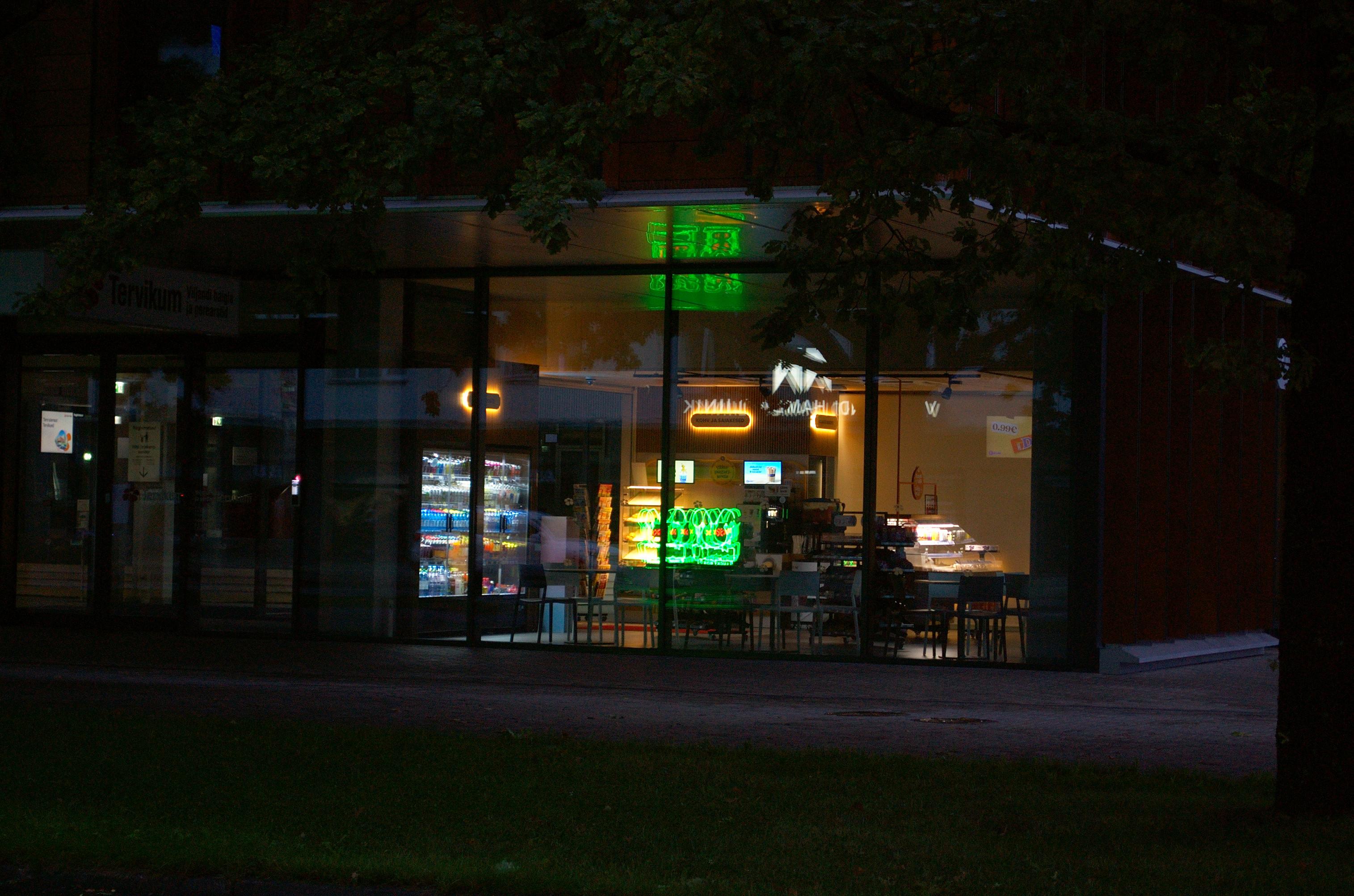Photo of a large display window of a cafe. The interior is dimly lit in yellow, showing dark silhouettes of tables and chairs. Along one wall there’s a refrigerator with glass doors, lit inside. A green neon sign across the street is reflected twice on the window.