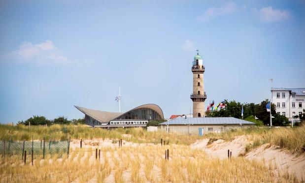 The image captures a serene coastal scene under a clear blue sky with scattered clouds. Dominating the right side is a tall, historic lighthouse with a green dome, surrounded by a cluster of colorful flags. To its left, a modern, angular building with a curved roof and large glass windows stands out, blending contemporary architecture with the natural surroundings. In the foreground, golden sand dunes stretch across the image, framed by sparse vegetation and a simple wooden fence.