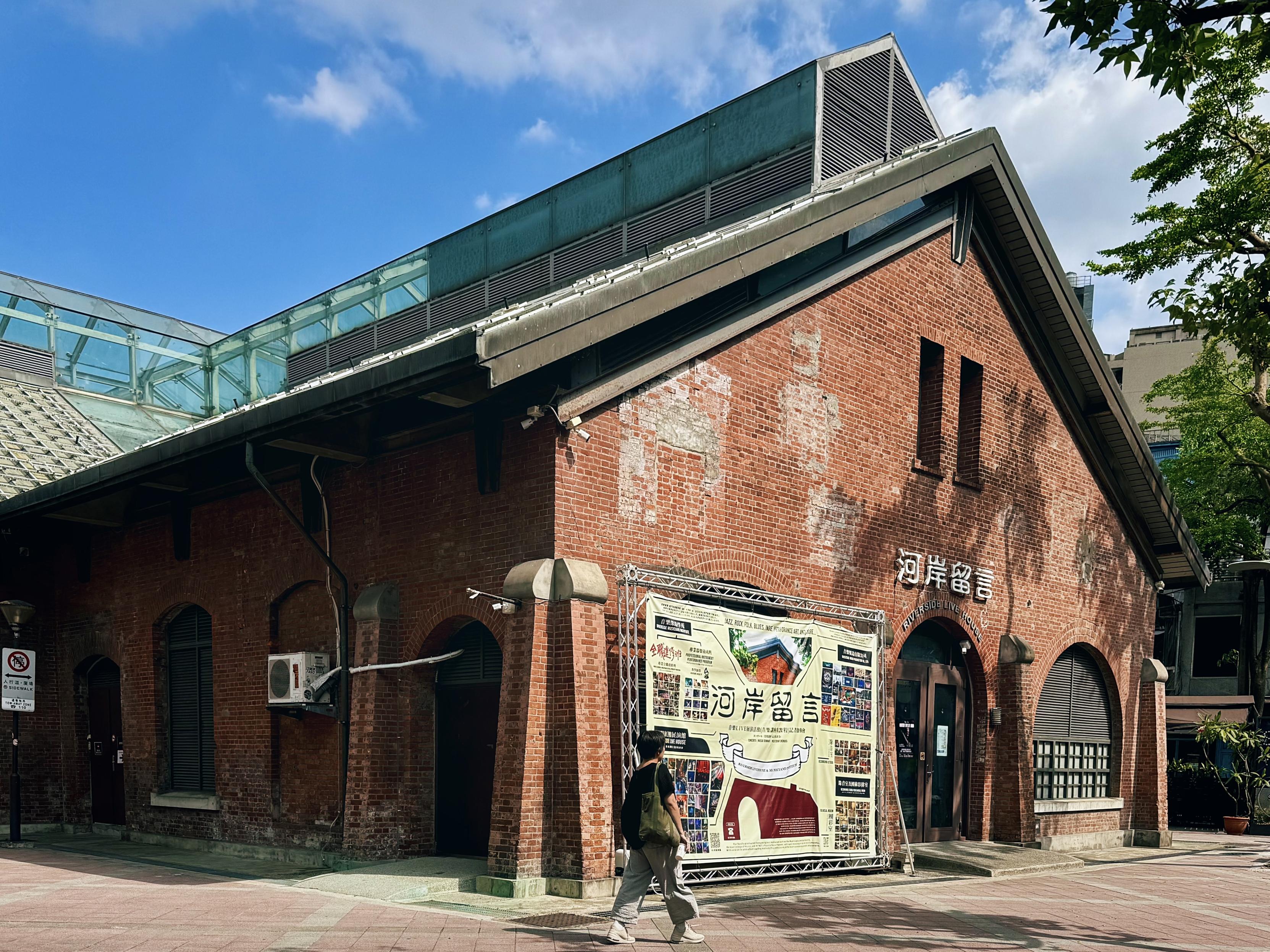 A person walks past a red brick building with arched windows and a sign in Asian characters, under a partly cloudy blue sky.