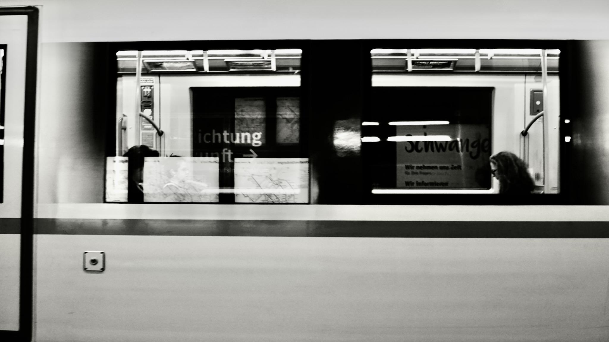 Picture of a subway train in Vienna, slightly moving. The wagon is bright with two big windows. It is nearly empty, on the right side is a single woman sitting at the window looking down. She has long hair and dark glasses. She is seen from the side.
Some reflections of the station can be noticed on the left side window. Picture is black and white.