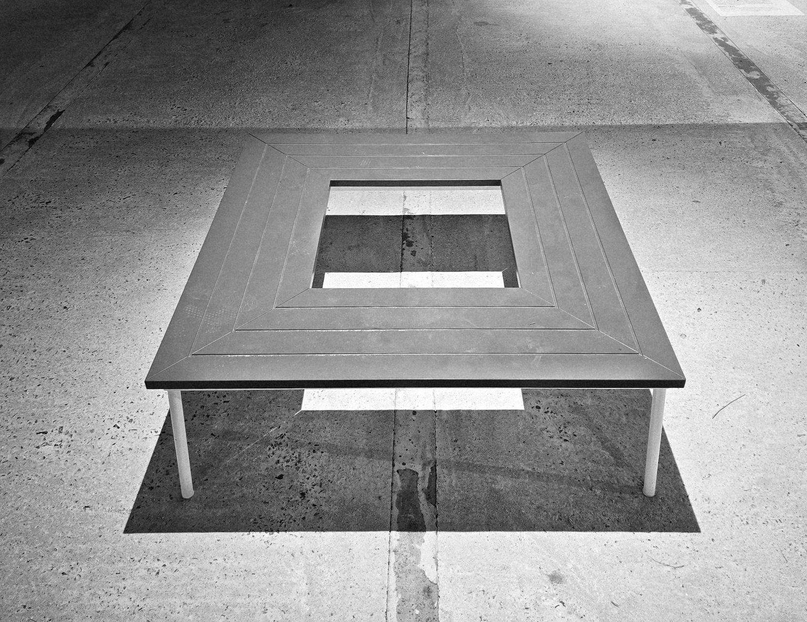 Schwarzweiß. Sitzbank quadratisch. Durch das harte Licht sind klare Linien und Schatten zu sehen.