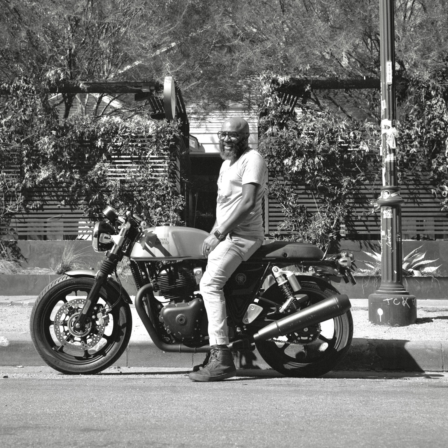 A black-and-white film photo. Reggie is seated on a Royal Enfield motorcycle in front of Daydreamer in East Austin, Texas. Monday, August 17, 2026.