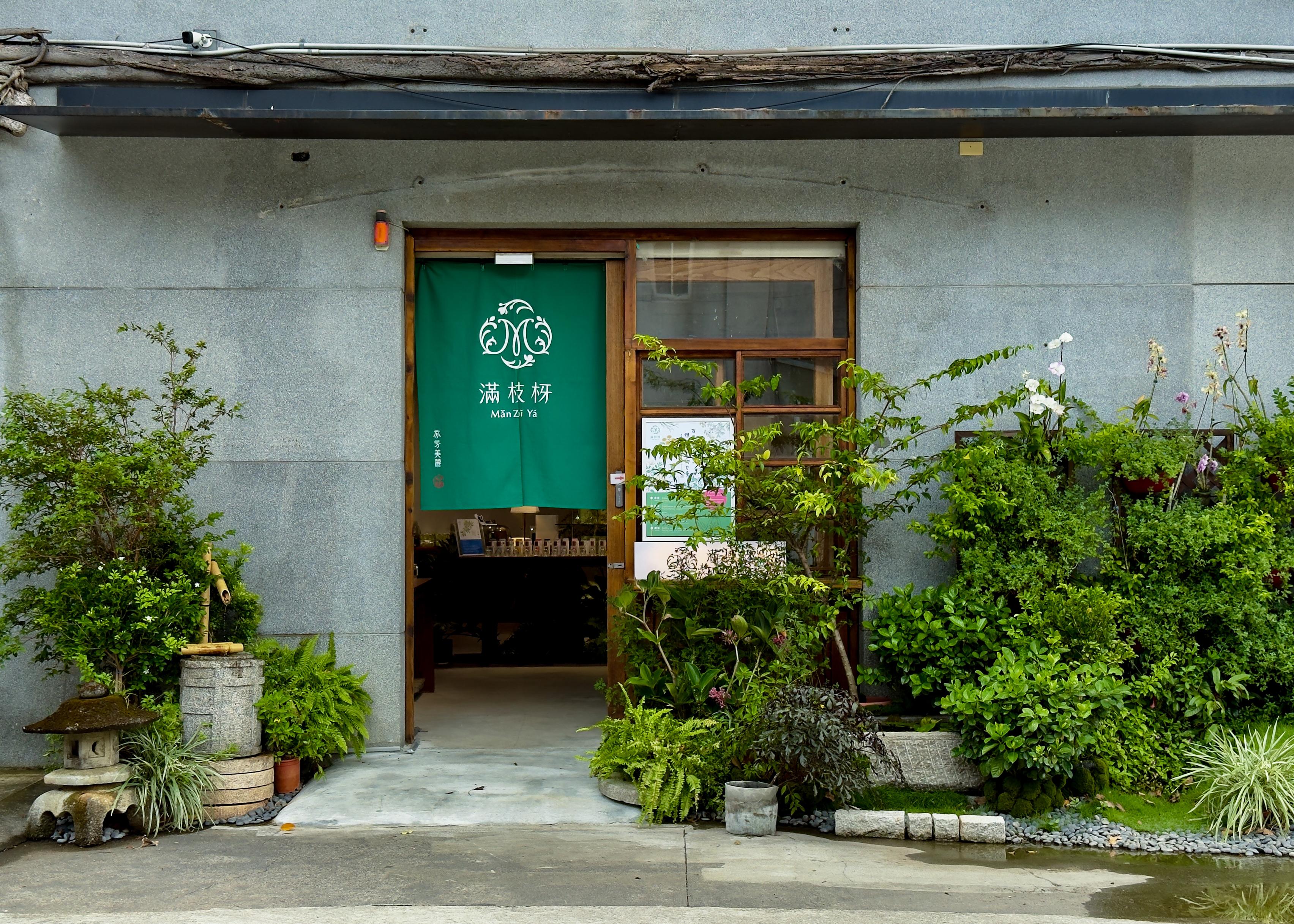 一家绿意盎然的日式庭院小店门口，有植物和石灯笼，门挂着绿色布帘，写有汉字和拼音。