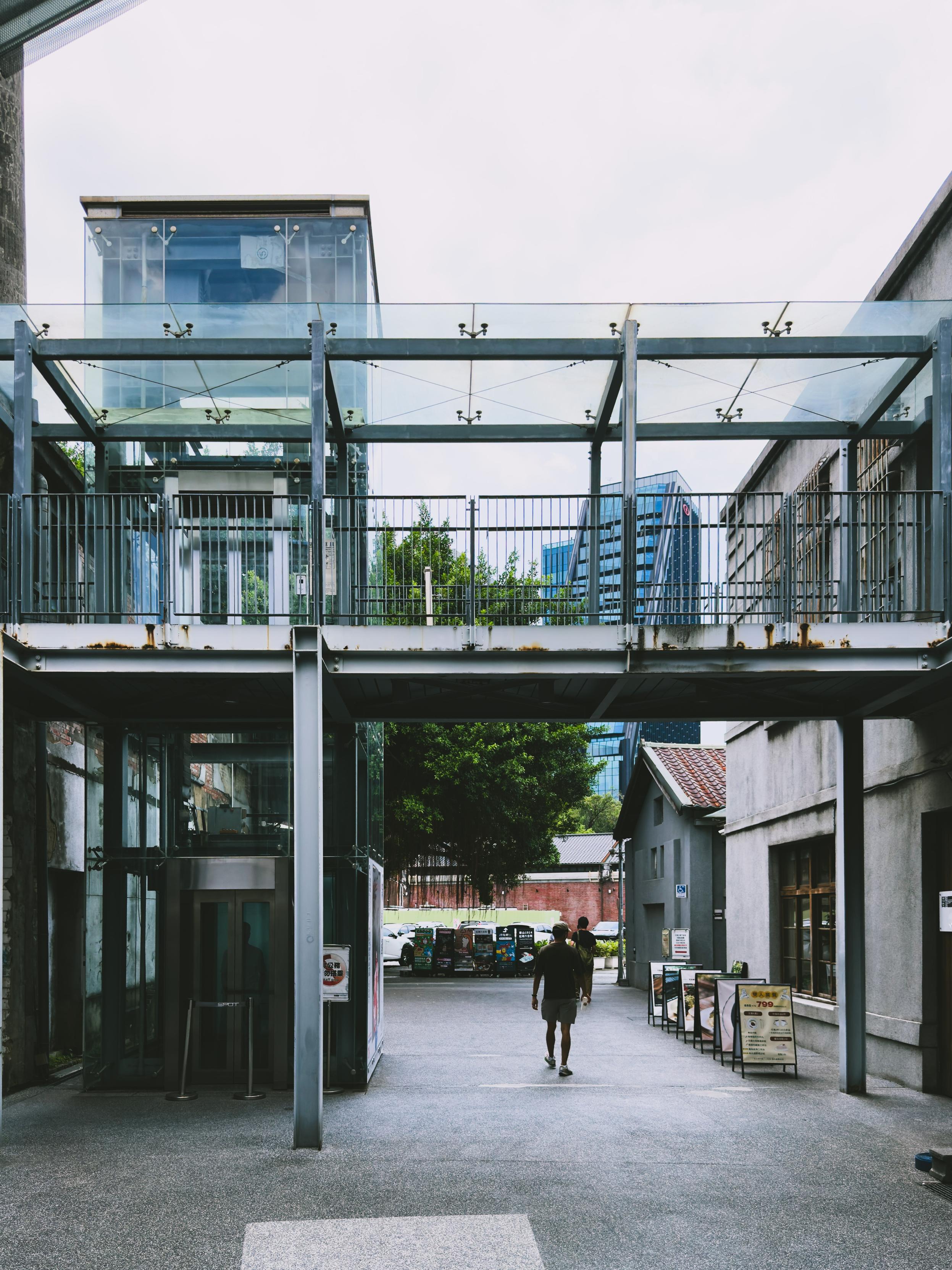A person walking through an urban passageway beneath a metal and glass structure, with a tree and modern buildings in the background.