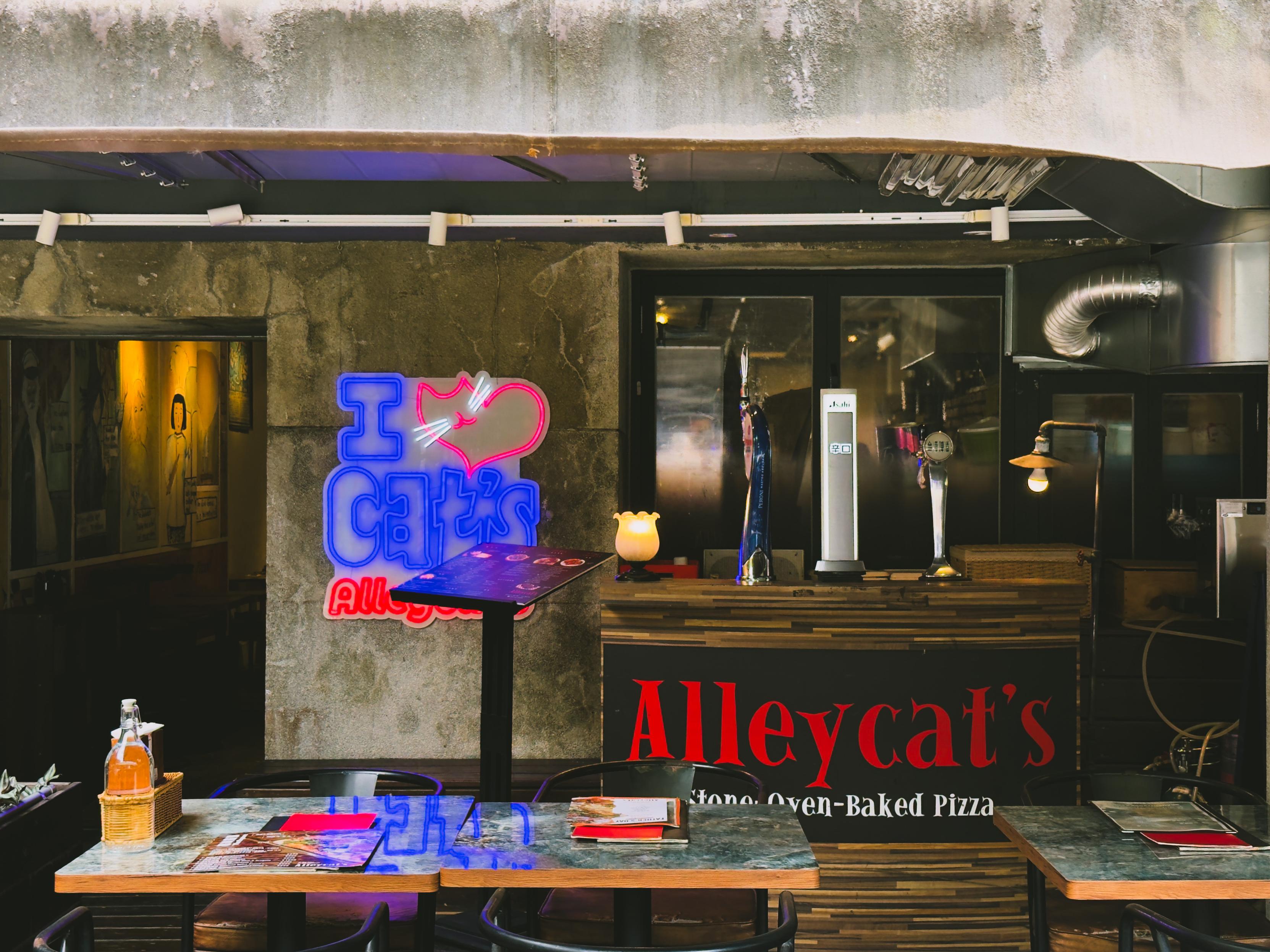 Neon sign reading "I ♥️ Cat's" at Alleycat's pizza restaurant, with tables and menus in the foreground.