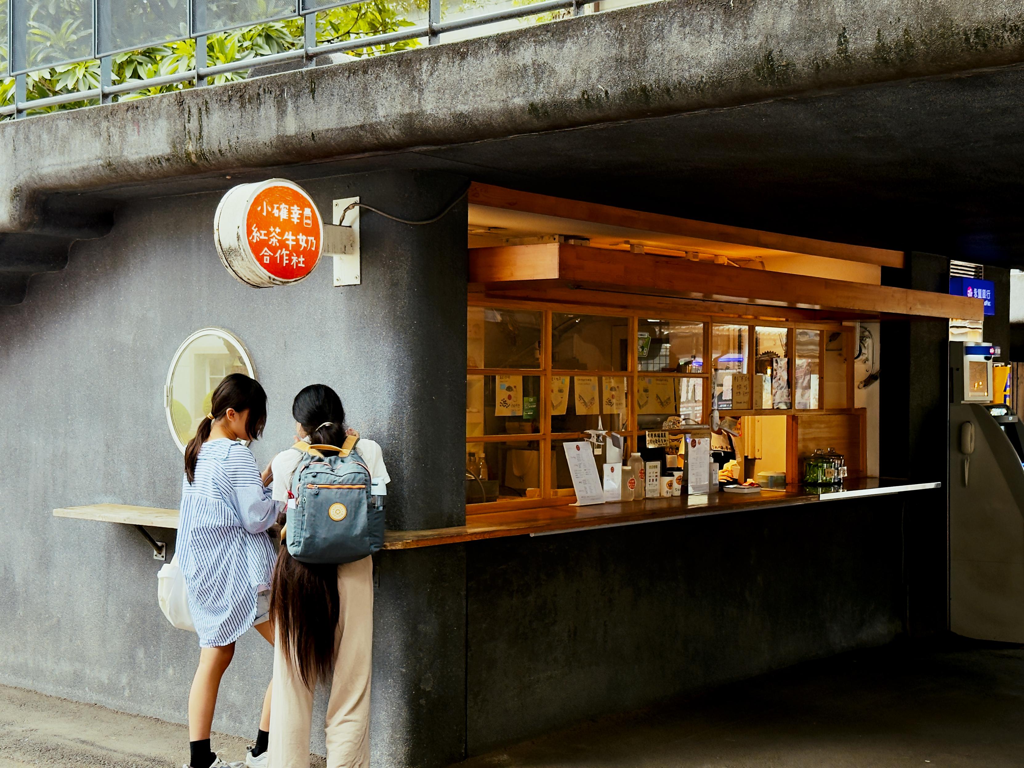 Two young girls with backpacks stand at a small outdoor food stand, looking at a menu or ordering food.