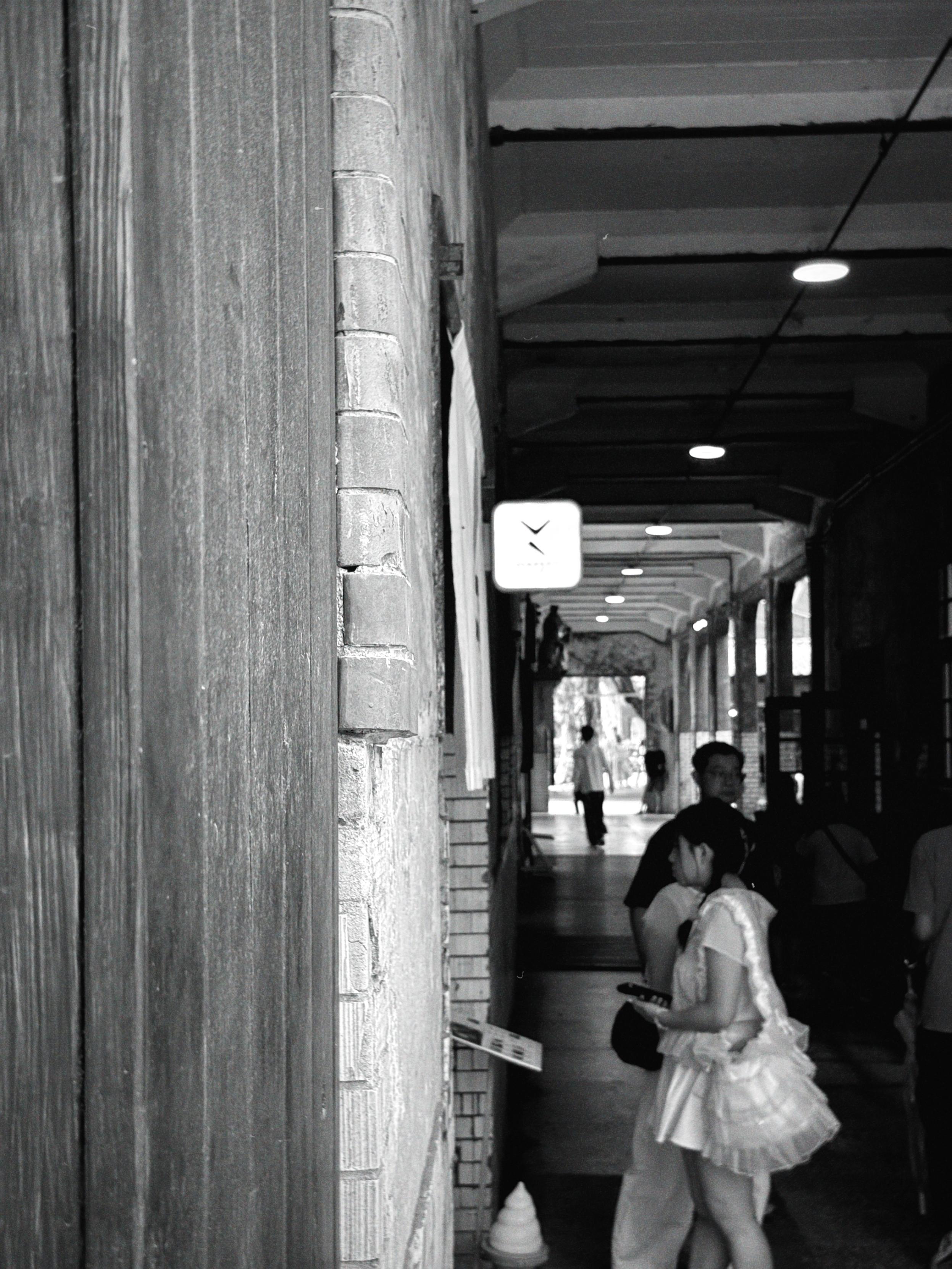 A black and white photo of a busy indoor corridor with people walking and sitting, with a brick wall on the left and ceiling lights overhead.