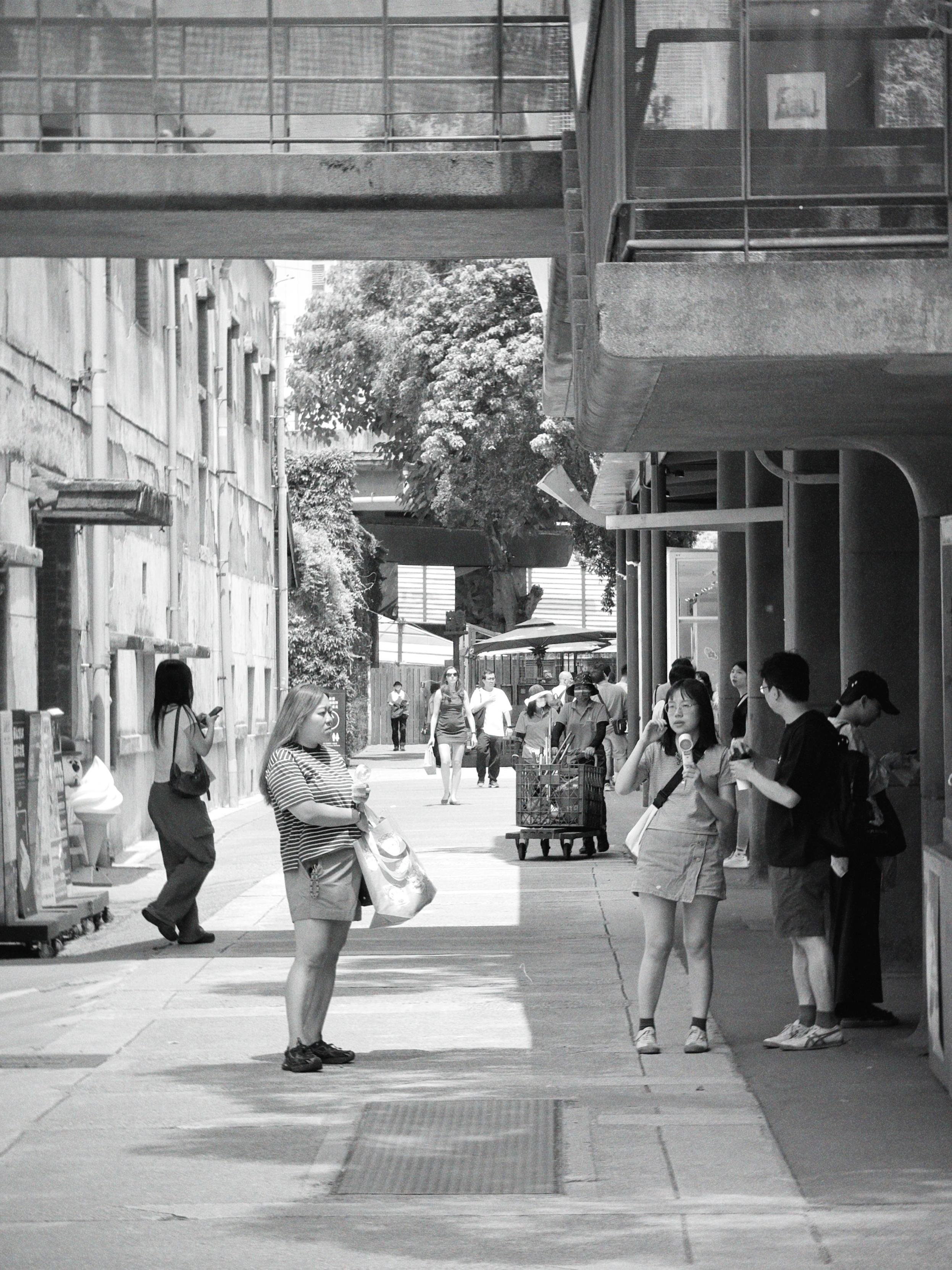 People walking and standing on a city sidewalk, some are engaged with their phones or food, with buildings and trees in the background.