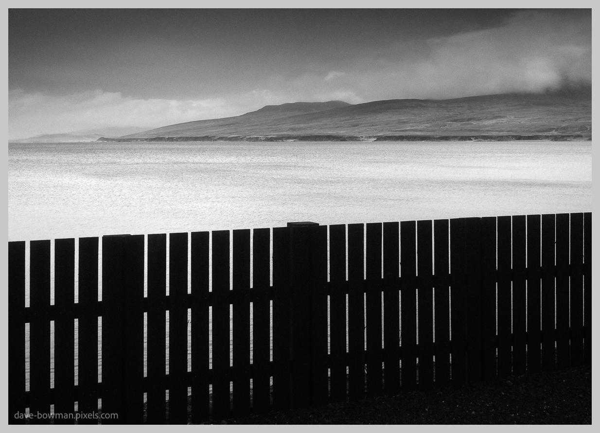 A photograph of a wide view across the Sound of Islay, with a simple wooden fence stretching across the foreground in dark silhouette. Beyond the fence, the calm water leads towards the distant, softly outlined hills of Jura, creating a strong sense of depth and separation between the nearby shoreline and the landscape on the far side. The entire scene is presented in black and white.