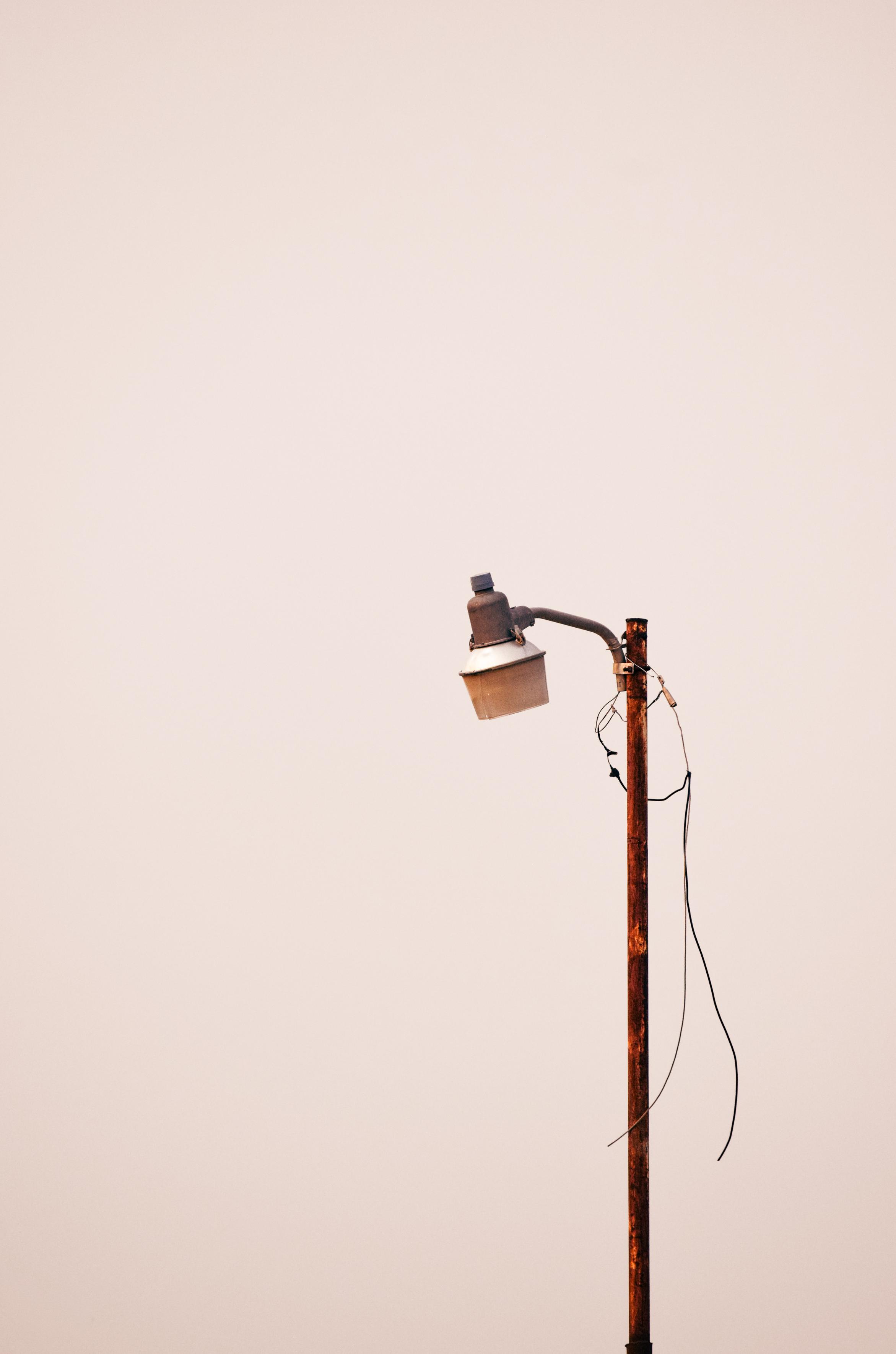 A weathered rusty utility pole with a tilted streetlamp and dangling wires stands alone against a pale pink sky, creating a quiet, lonely mood.