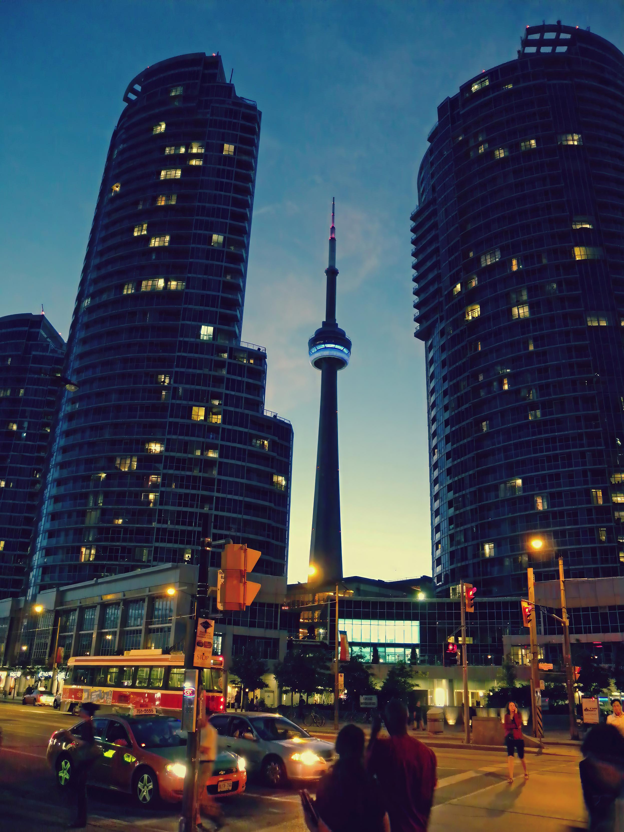 The photo shows the CN Tower in Toronto, picturesquely framed at dusk by two modern skyscrapers at the intersection of Queen’s Quay and York Street. Landmark: The 553-meter-tall television tower is Toronto’s most famous landmark and was, for a long time, the tallest freestanding tower in the world. Perspective: The photo looks north from the waterfront (Harborfront), where closely spaced residential and office complexes frame the view of the tower.