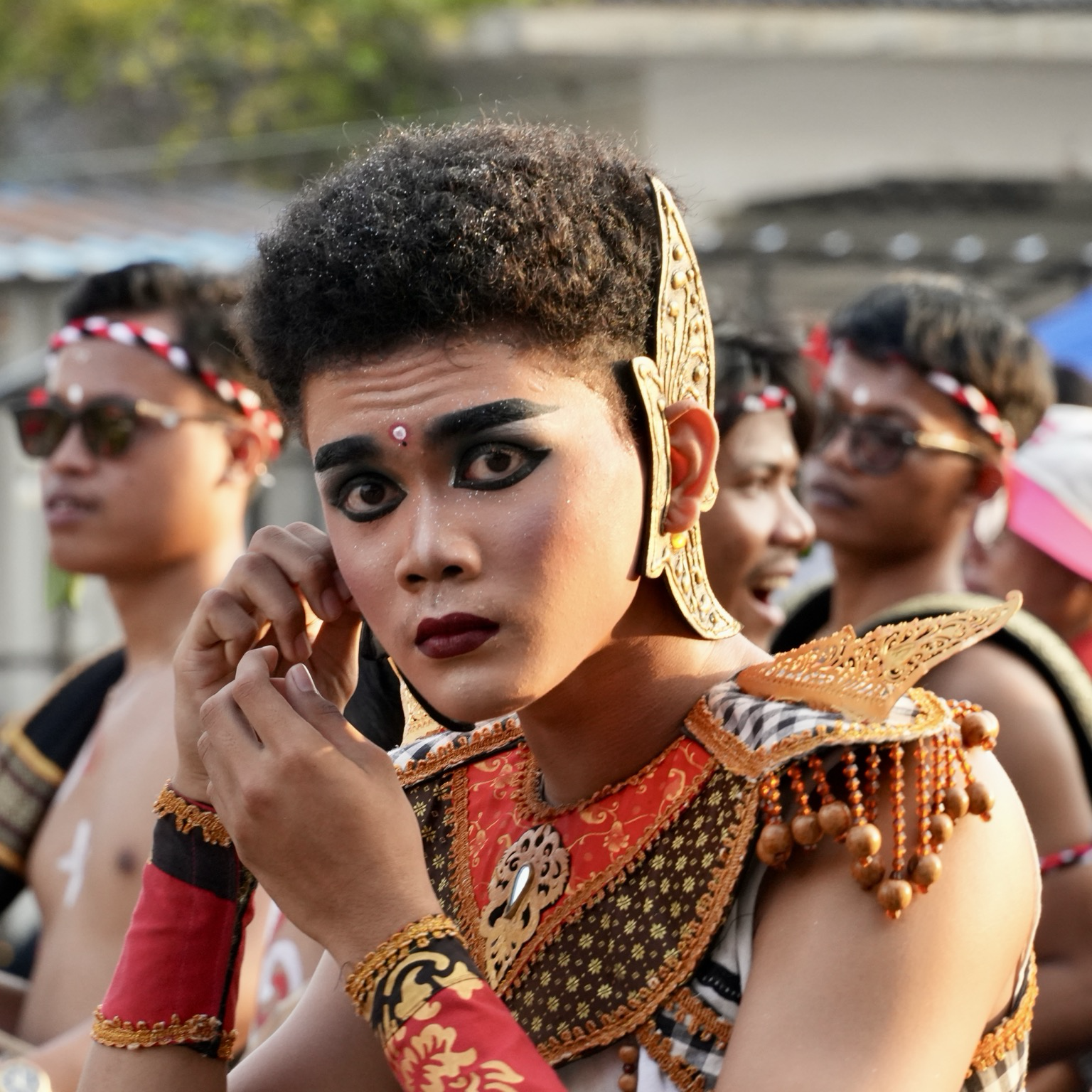 Close portrait of a young parade performer in an ornate red, black and gold costume, adjusting something by their ear while looking directly into the camera. Warm late-afternoon sunlight falls across the face, with other parade participants softly blurred in the background.