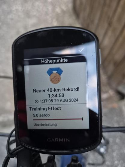 This image shows a **Garmin cycling computer** mounted on a bicycle handlebar. The screen displays a notification for a new 40-kilometer record achieved on August 29, 2024, at 1:34:53. The notification includes details such as "Training Effect" with a value of 5.0 aerobic and a label "Überlastung" (overload in German). The device also shows a badge icon, likely representing the achievement.

