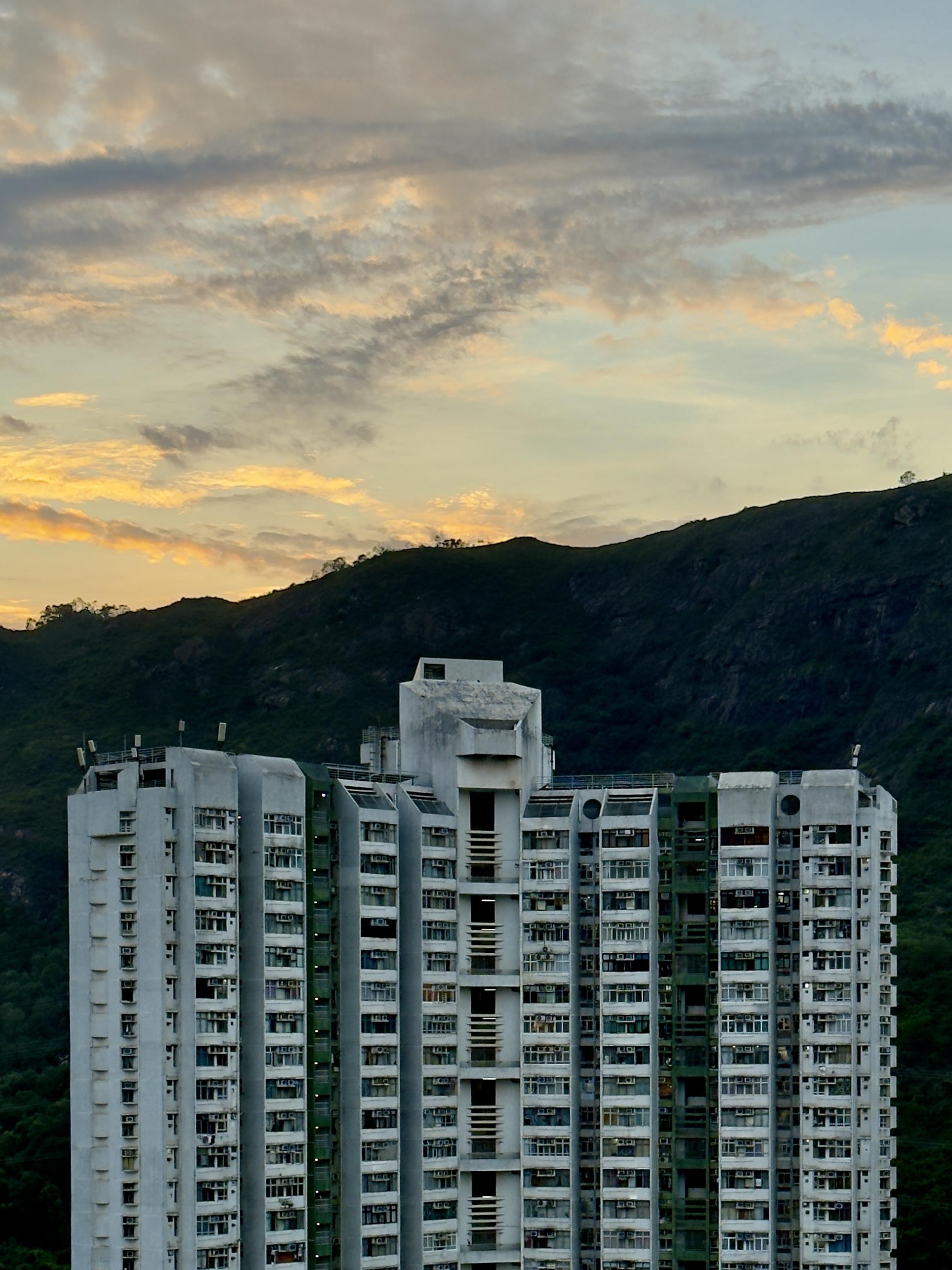 A tall white apartment building with many windows stands against a green mountain under a cloudy, partly sunset sky.