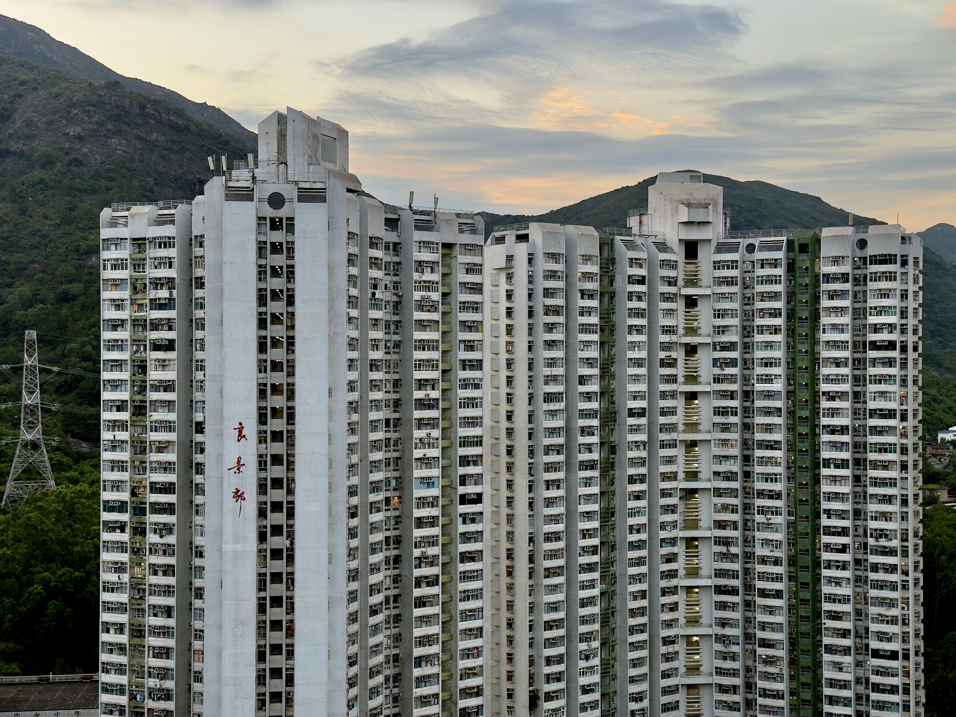 一栋高层住宅楼，背靠山脉，晚霞映照，建筑外墙上有红色汉字。