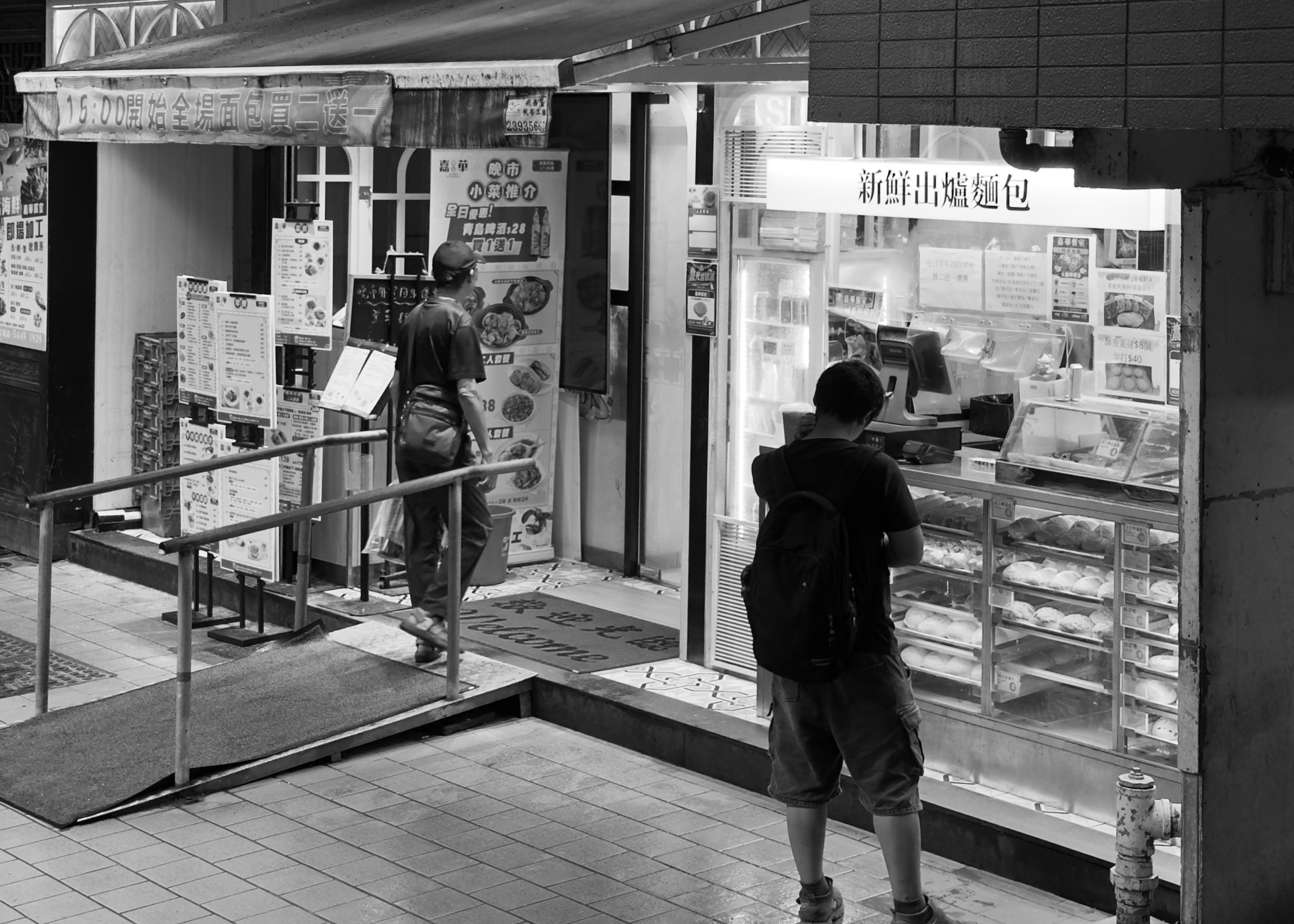 两人站在街边的面包店前，一人正在付款，一人等待购买。