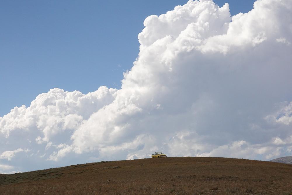 a broad, open hill top with a yellow truck, under huge, billowing white clouds in a blue sky