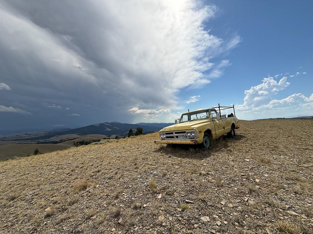 A broken down old yellow pick up truck sits on a rocky hilltop under a blue sky with dramatic clouds