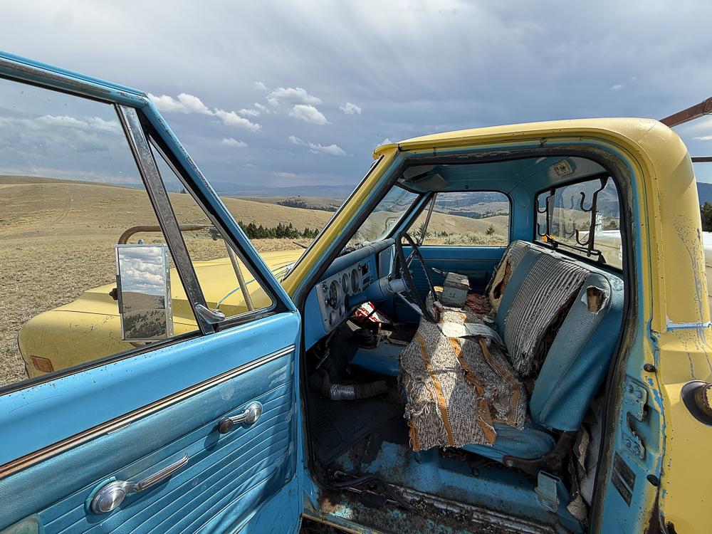 an old yellow pick up truck with a blue interior which has been chewed on, stands with its door open, reflecting the view behind the camera, in the door mirror