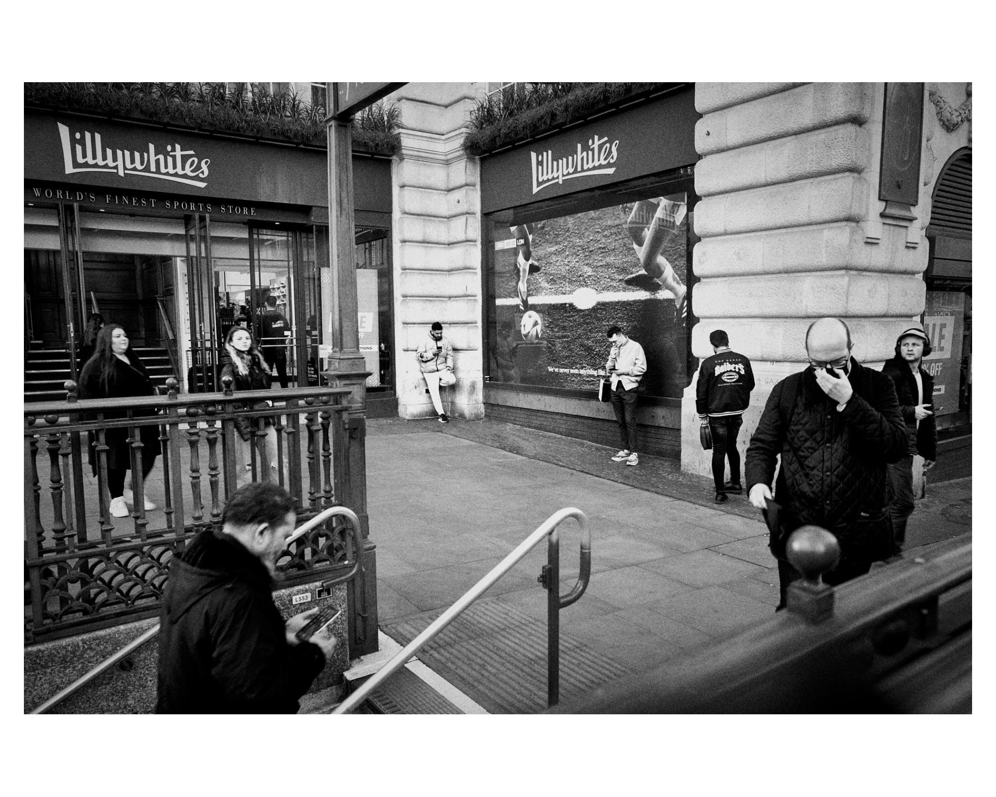 A corner in London, the tube, people distracted, on phones, etc. spacing dividing. Alone but together. black and white photo. street photography