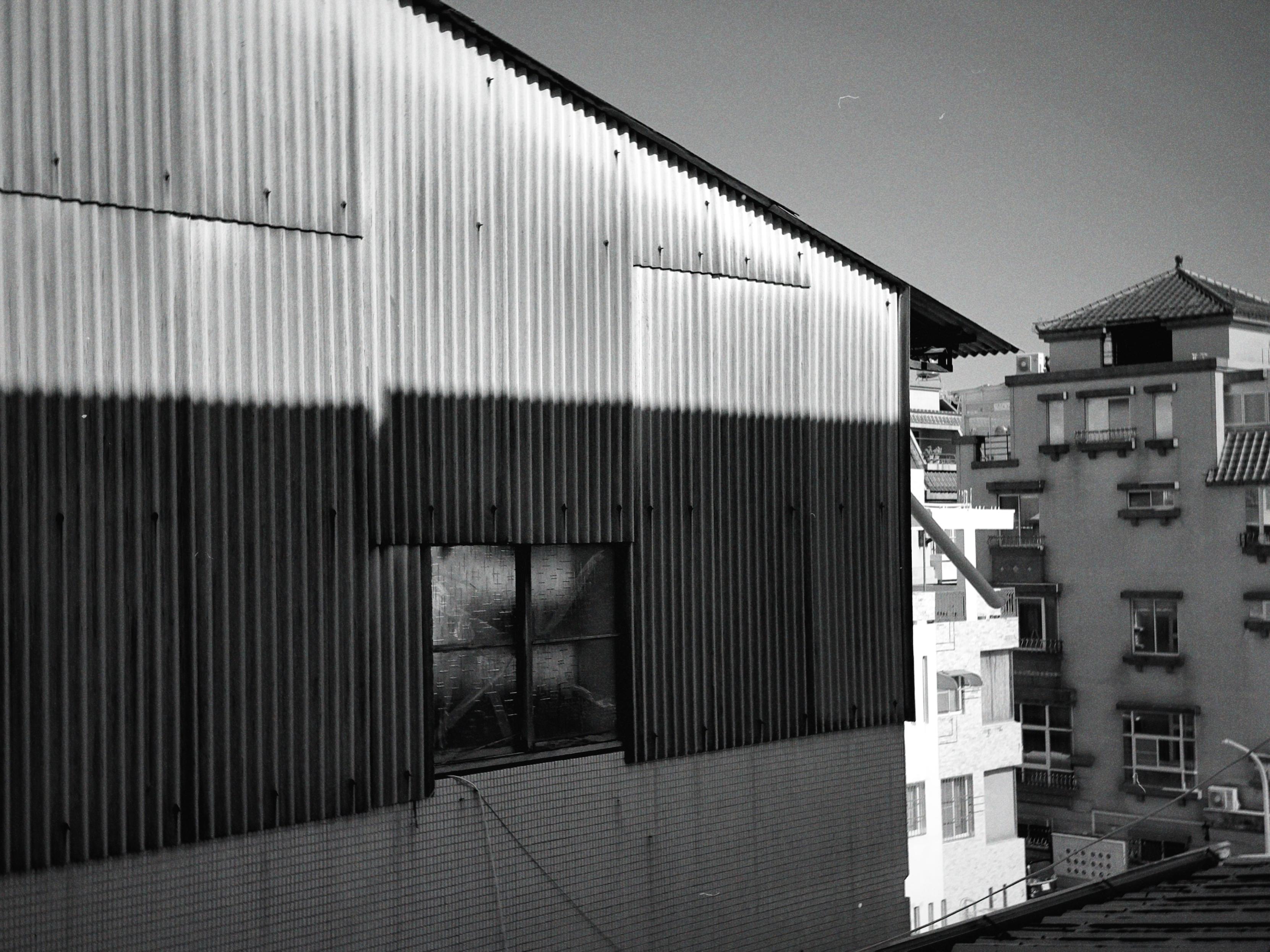 A black and white photo of buildings, with shadows cast on the nearby corrugated metal wall, under a clear sky.