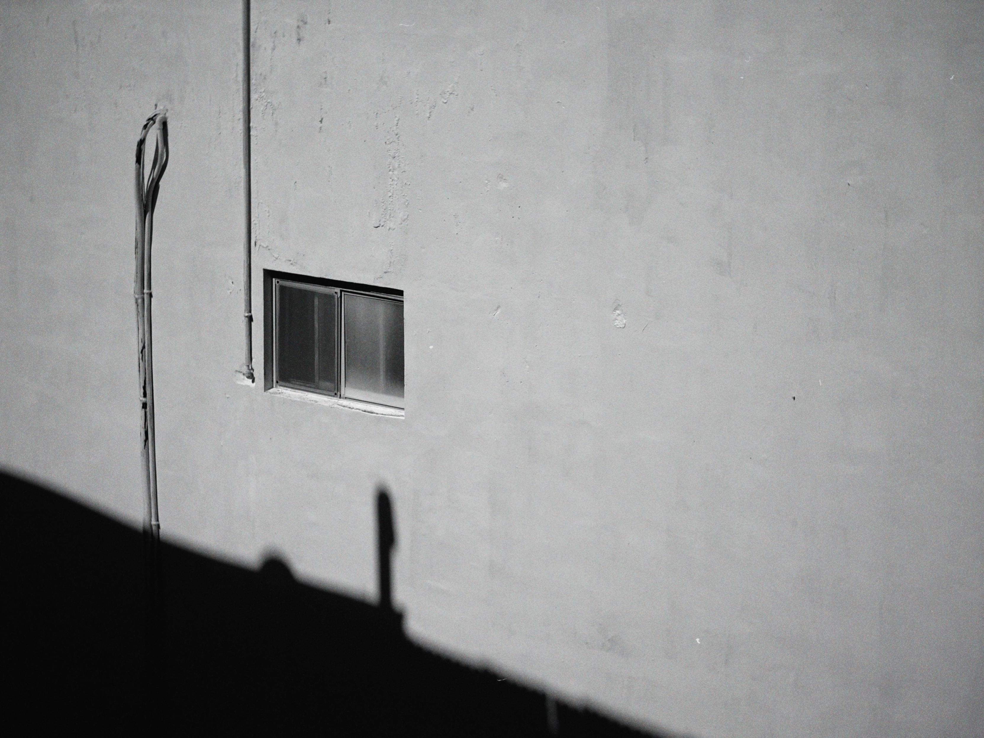 A small window in a plain white wall with some visible texture and an exposed pipe running vertically on the left side, casting a shadow.