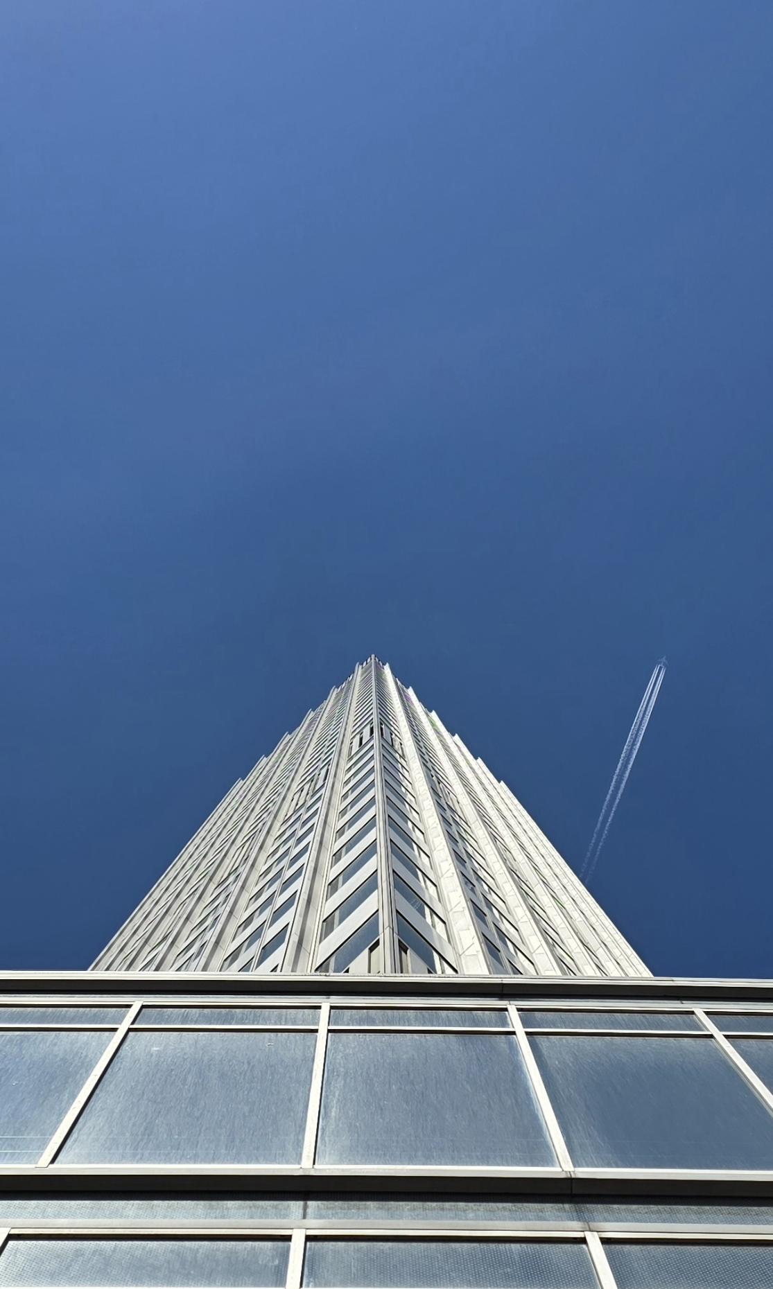 Silbergraues Hochhaus berührt den blauen Himmel. Ein Flugzeug schwebt durchs Bild.
