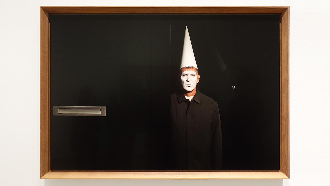 Een foto uit de serie April Fool uit 2020. Tegen die tijd dat de gezondheid van Erwin Olaf hard achteruit. Erwin presenteert zich als joker, als mimespeler en laat ons onbewogen kennismaken met zijn diepste onrust. In dit zwarte schilderij staat Erwin Olaf met een wit geschminkt gezicht en een witte puntmuts ons aan te kijken in een deuropening. Alleen de brievenbus en het kijkgat in de deur zijn zichtbaar. Komen we binnen in zijn leven? Willen wij wel binnen komen in zijn leven? Met alles waar hij zich zorgen over maakt. Met zijn strijd tegen zijn geaardheid? Met onze koopdrift? Met echt concentreren wat belangrijk is?