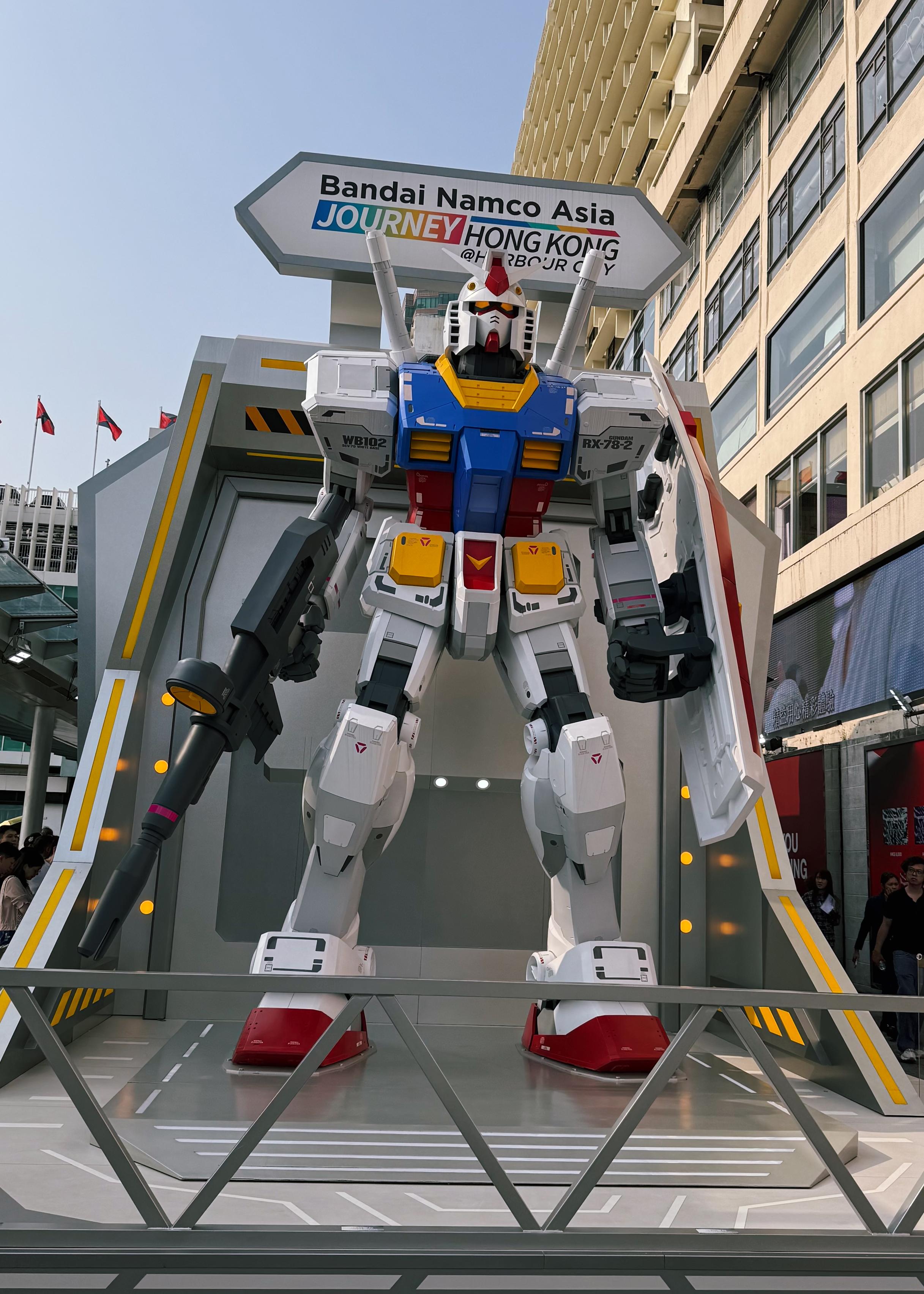 高达模型展示在户外，背景为繁忙的市区街道和建筑，标志上写有“Bandai Namco Asia”等字样。