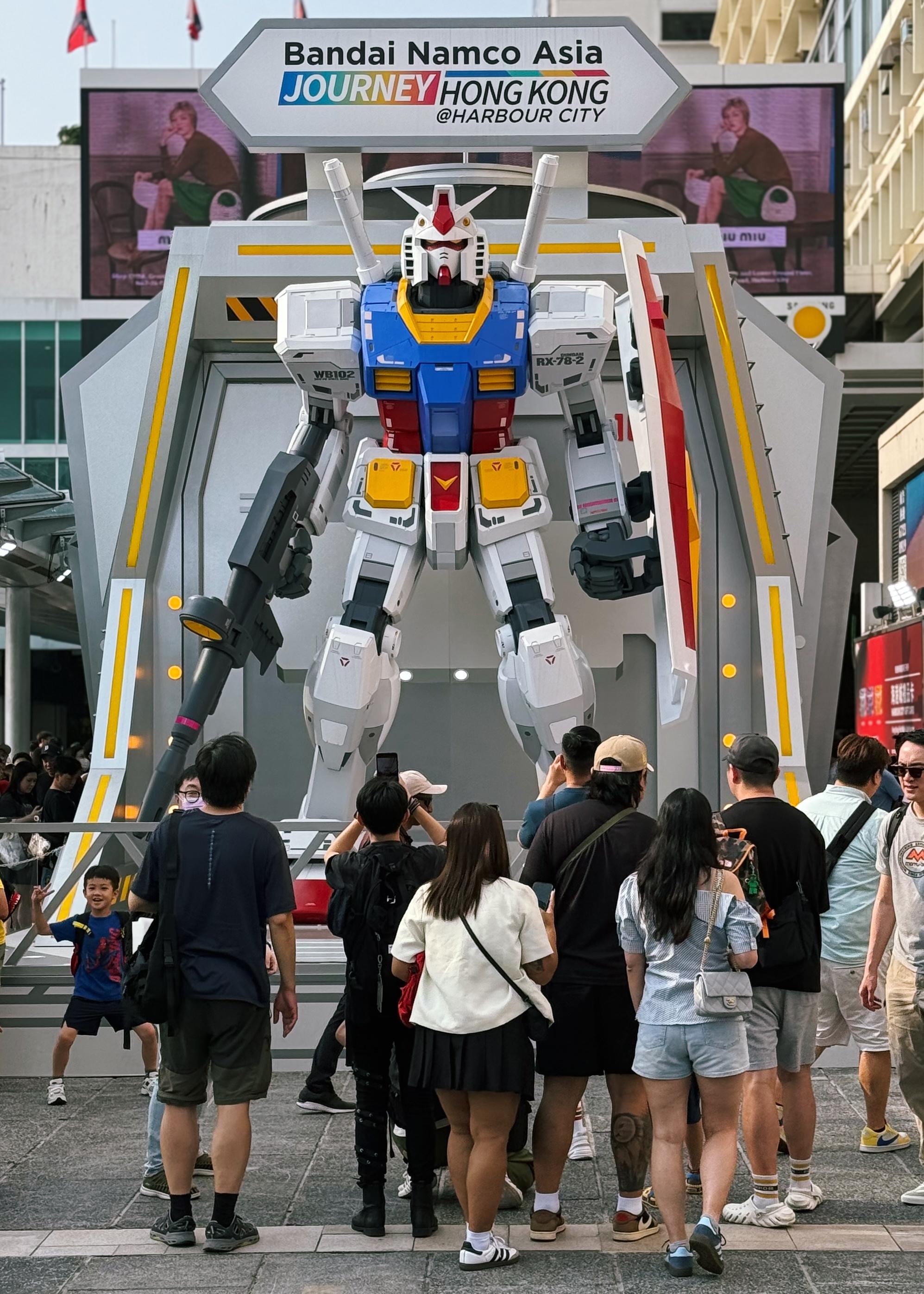 高达模型展览，众人在模型前拍照，背景为“Bandai Namco Asia”横幅和电子屏幕。