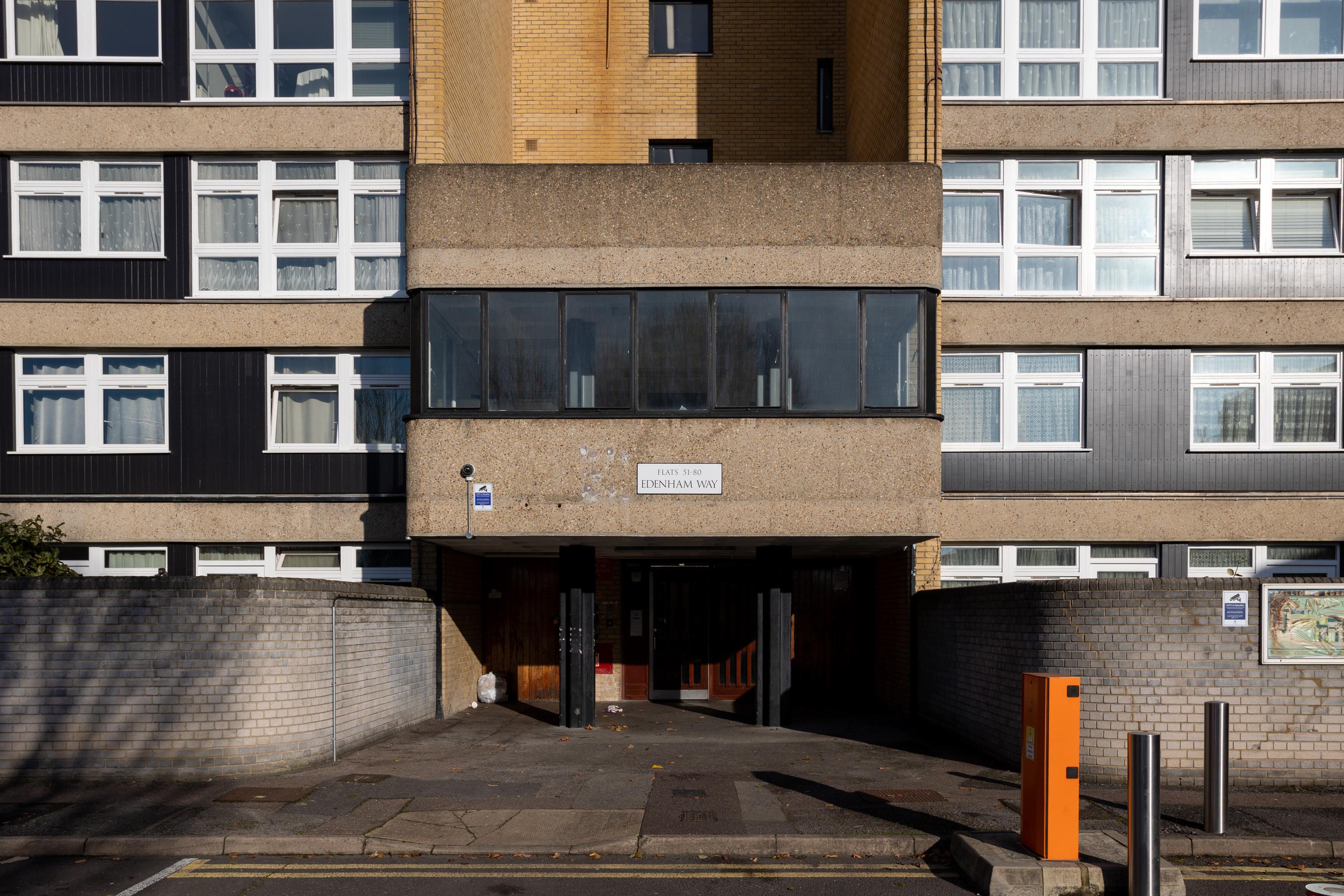 A modern block of flats entrance with a concrete overhang, large windows above, brick and grey panel facade, and a sign reading Denman Wu. A gated entry, some litter, and an orange parking barrier are visible in the foreground.