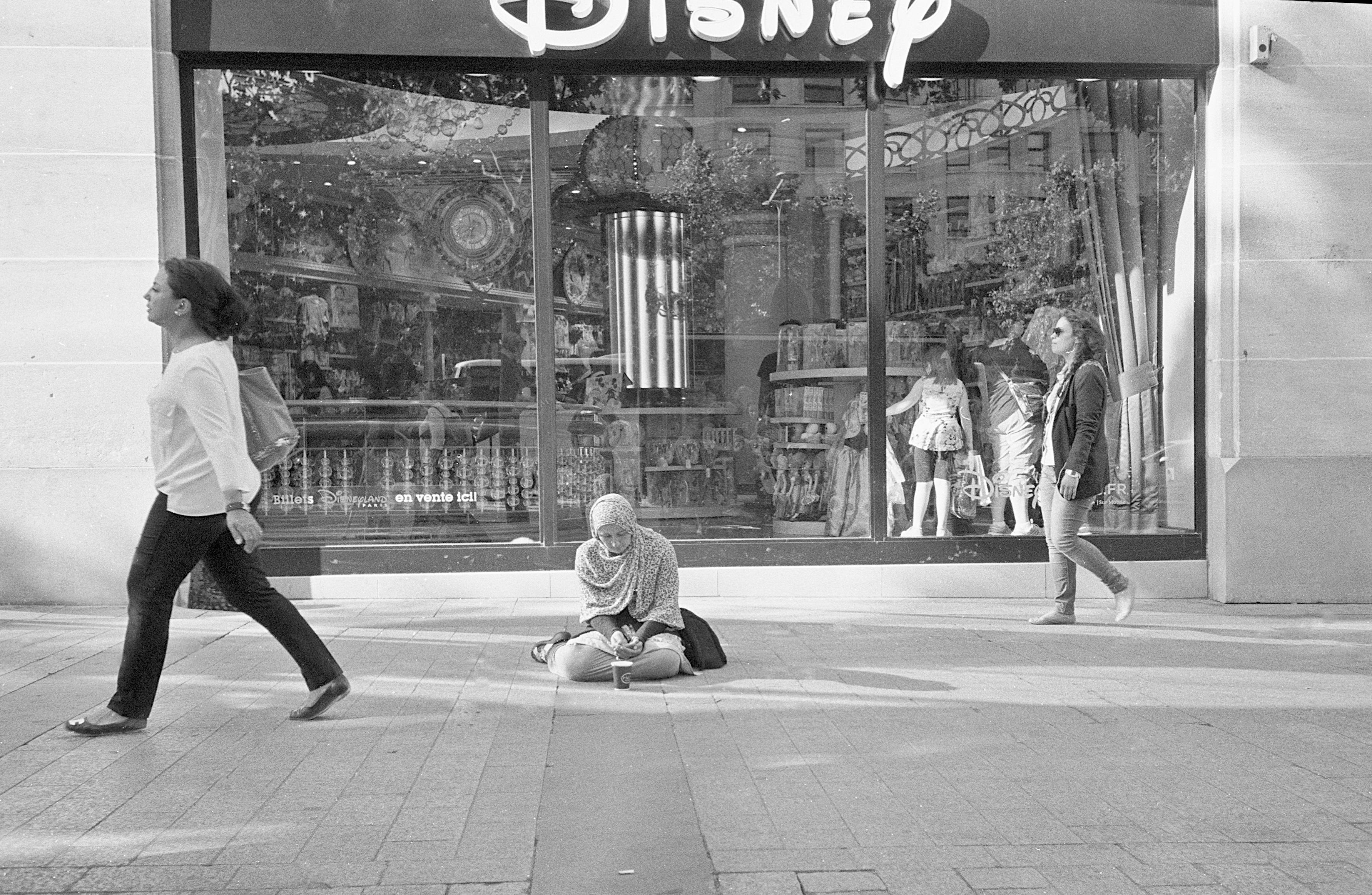 une femme mendiant agenouillée devant une vitrine Disney, deux passantes l'ignorent