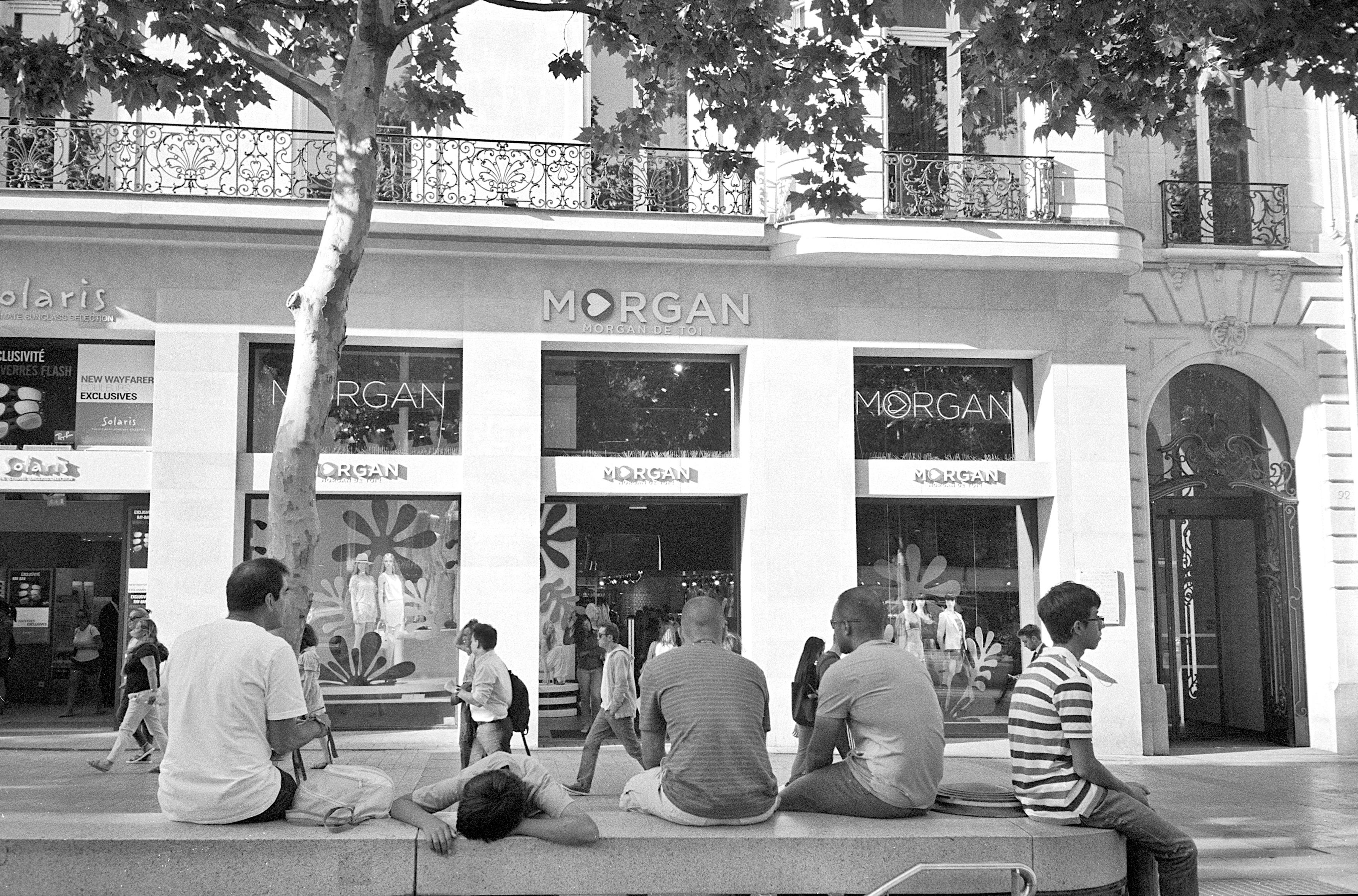 des personnes assises sur un muret, de dos, face au magasin Morgan des Champs Elysees. Une des personnes est avachie, endormie.