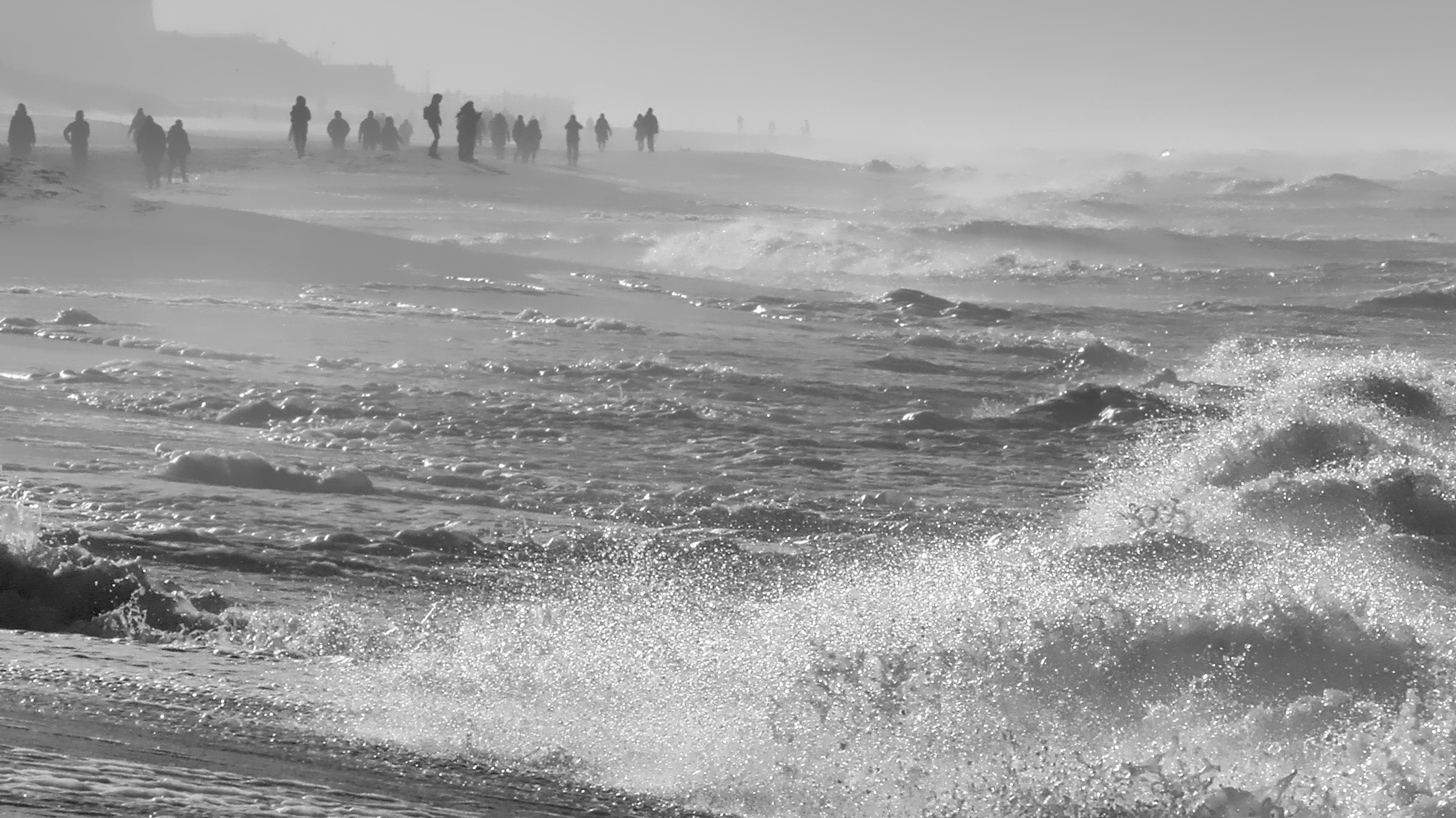 Schwarzweiß-Foto eines Strandes an der Nordseeküste bei stürmischem Seegang und langer Brandung an den Strand. Einige Menschen beobachten die Wellen und sind im Gegenlicht nur als Silhouetten zu sehen.