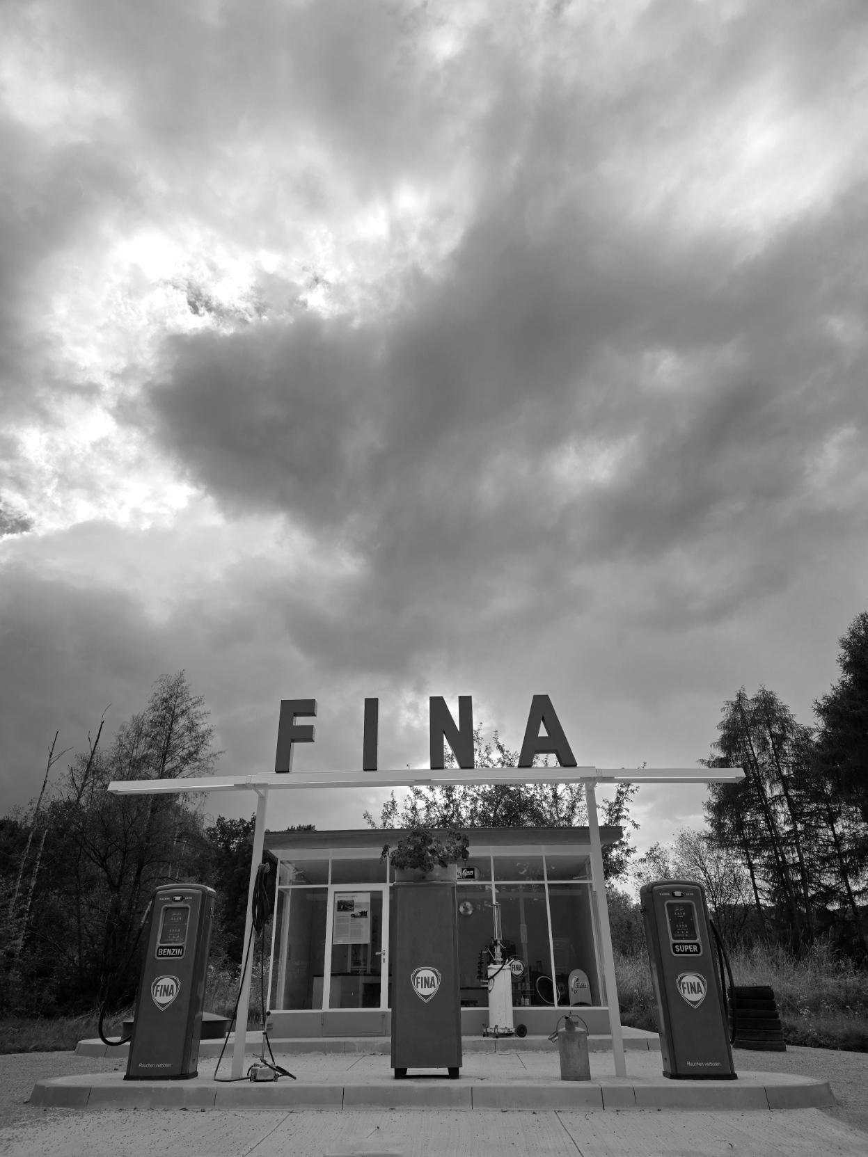 Black and white. A gas station from 1959. The word “Fina” in the center. Above it, clouds, some with rain.