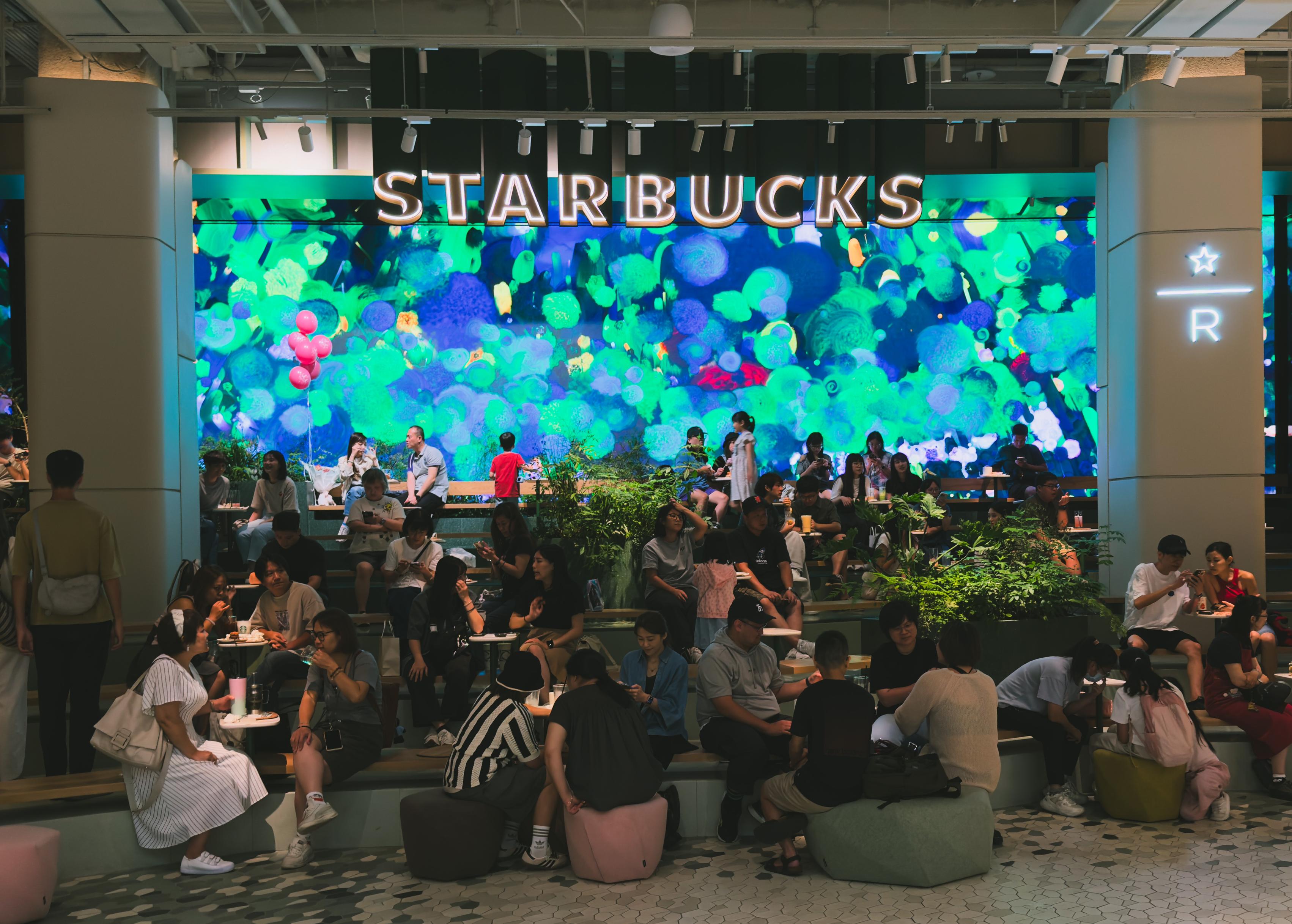 星巴克门店内，许多顾客在座位上享受咖啡，背景是一个色彩斑斓的数字屏幕。
