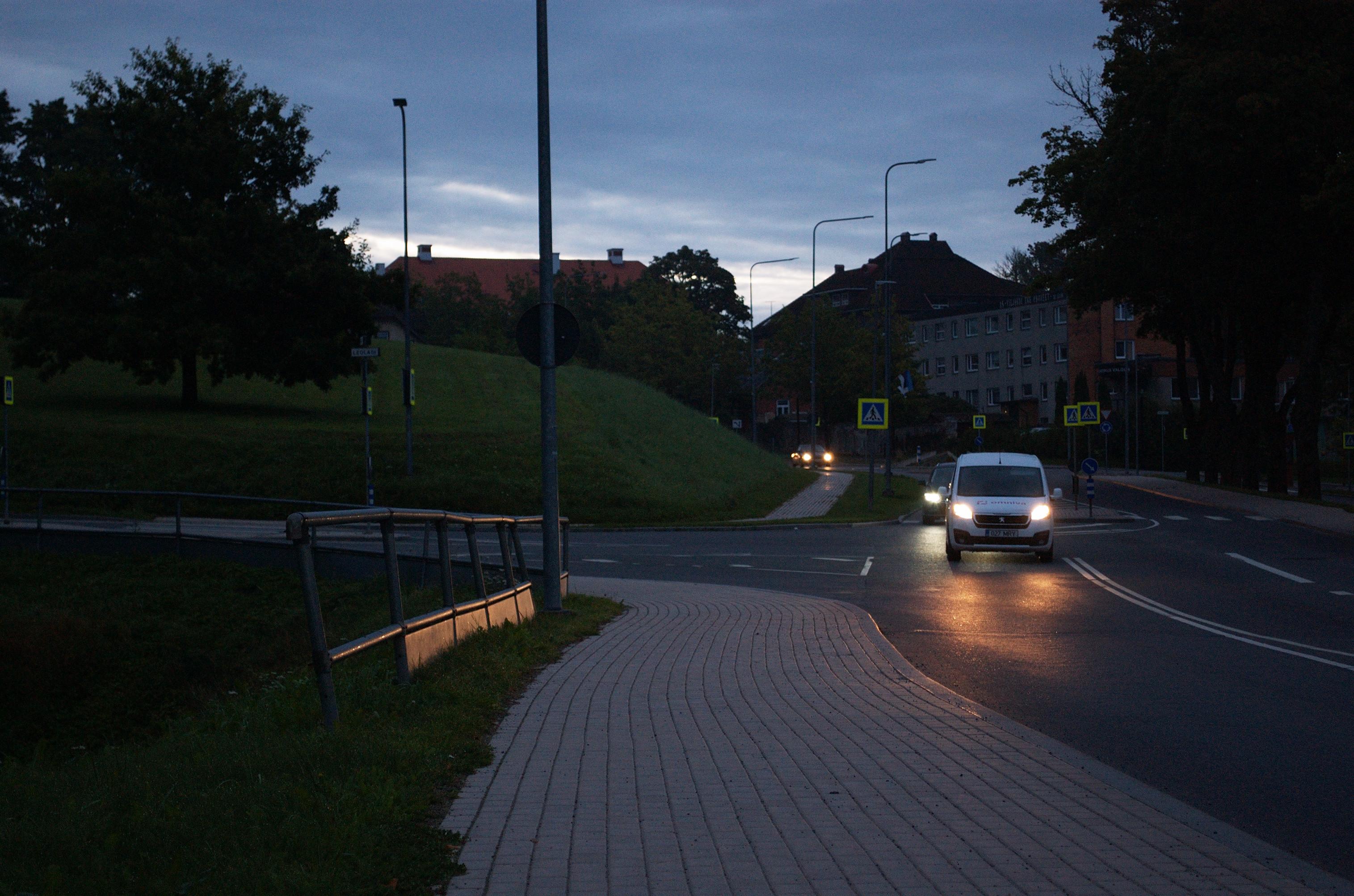 Early morning photo of a street running through a mostly empty part of the town. I’m standing on the sidewalk. To my left is a steel safety barrier blocking off a steep embankment. The oncoming cars’ headlights cast a yellow glare on the asphalt and reflect off the lower, solid part of the barrier.