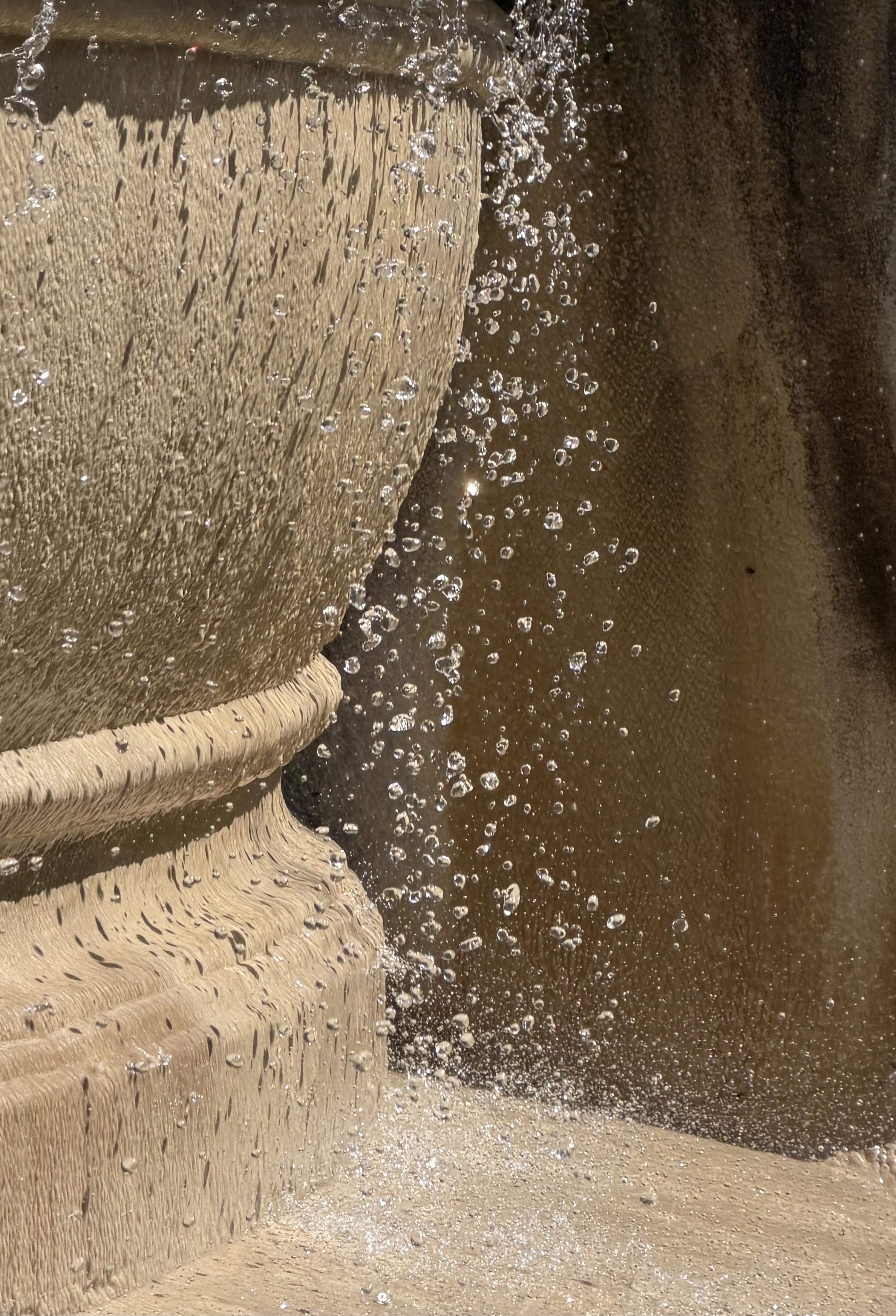 Water droplets suspended beside the curved stone form of a fountain, catching sunlight against a dark brown background.