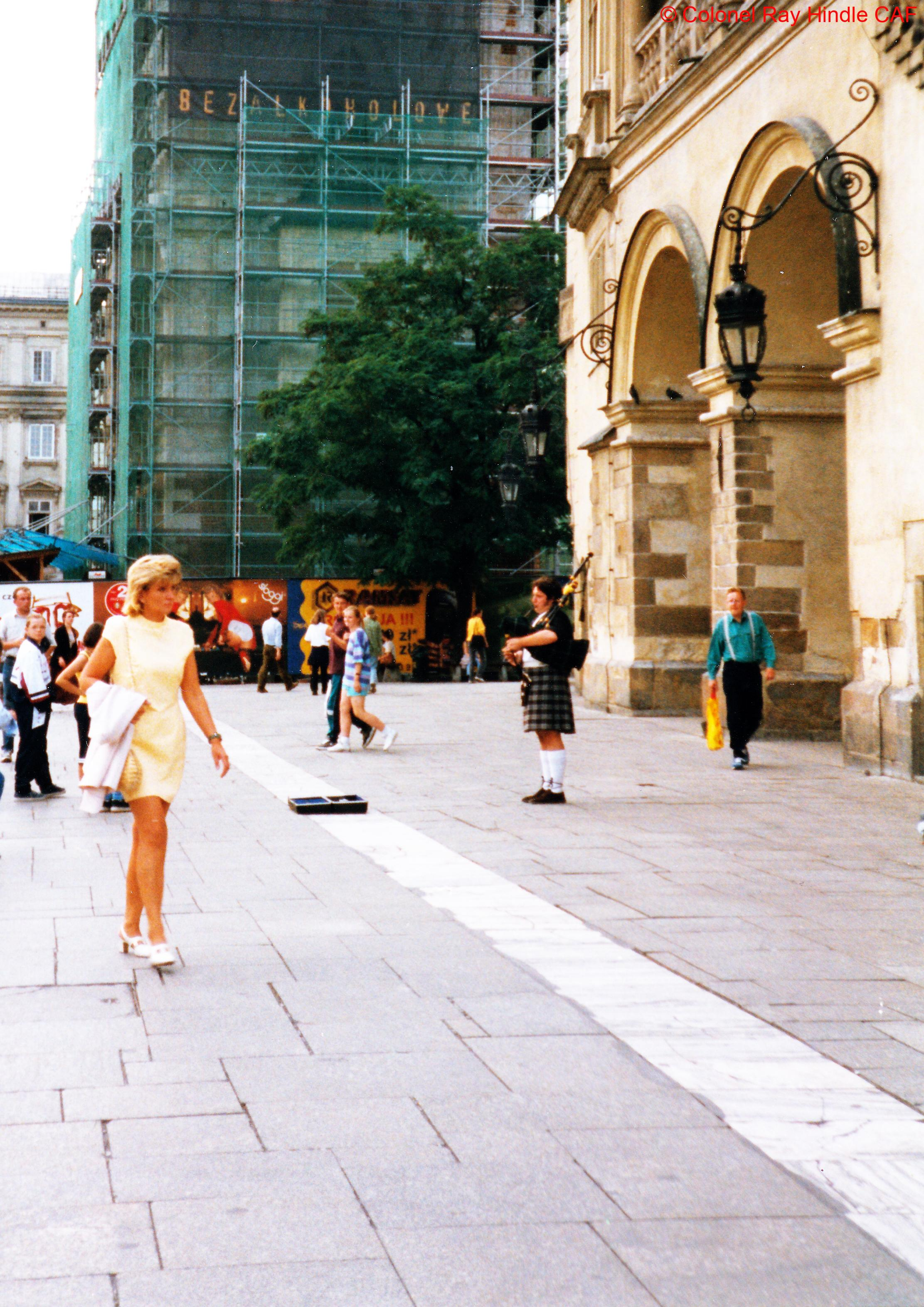 A Scots Piper plays in a busy pedestrian square with people walking past historic stone arcades and ornate black lanterns. In the background, a large building is covered in green construction scaffolding, with trees and colorful hoardings along the street. A woman in a light dress walks in the foreground, while other pedestrians gather and move through the plaza.