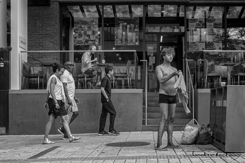 A man wearing underwear and sandals, is holding a piece of clothing after undressing on the sidewalk, in front of people passing by.