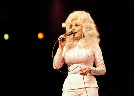 Dollar Parton sings into microphone with big hair and a few lights against a dark background