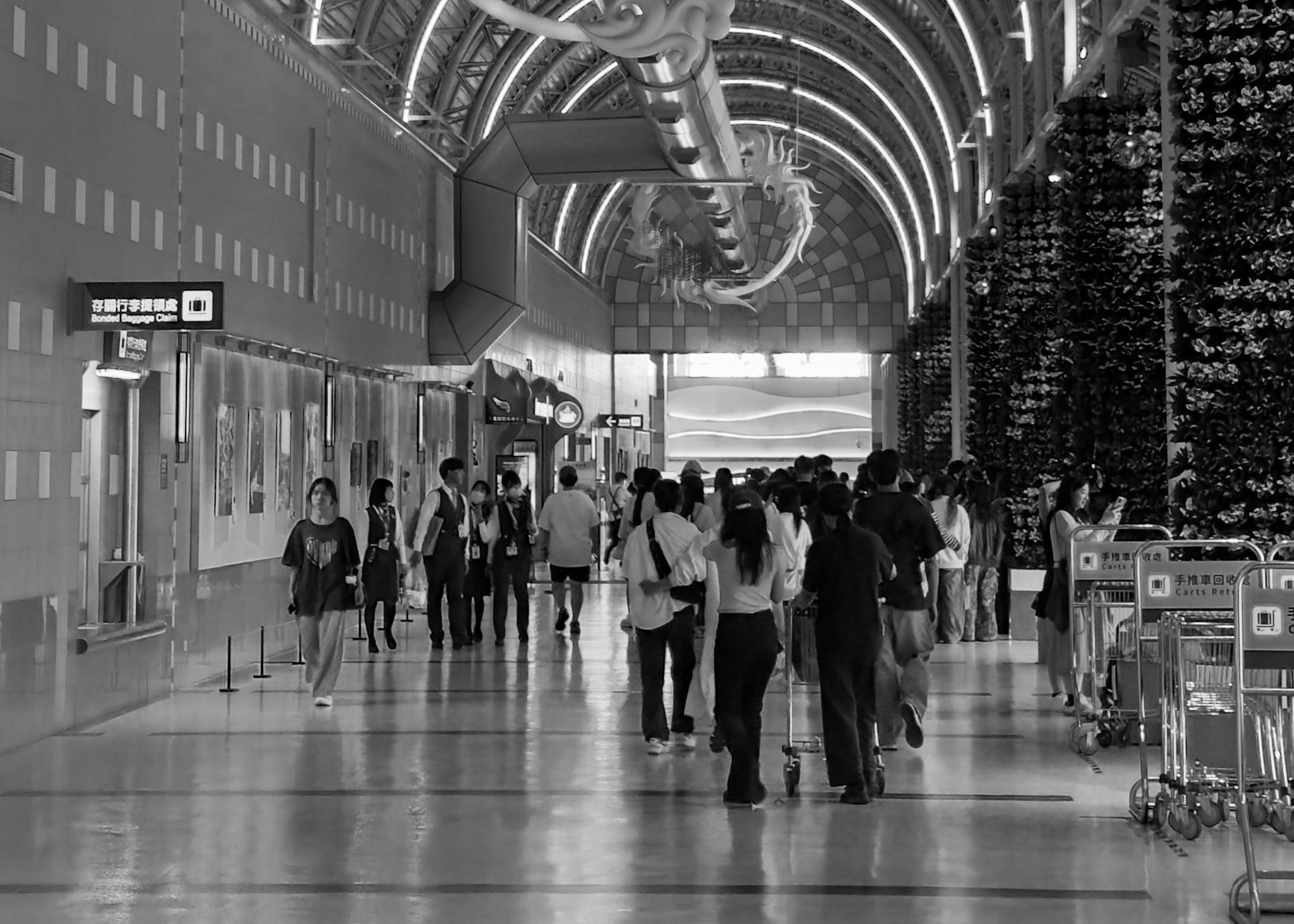 People walking and waiting in an airport terminal with high, arched ceilings and decorative lighting.
